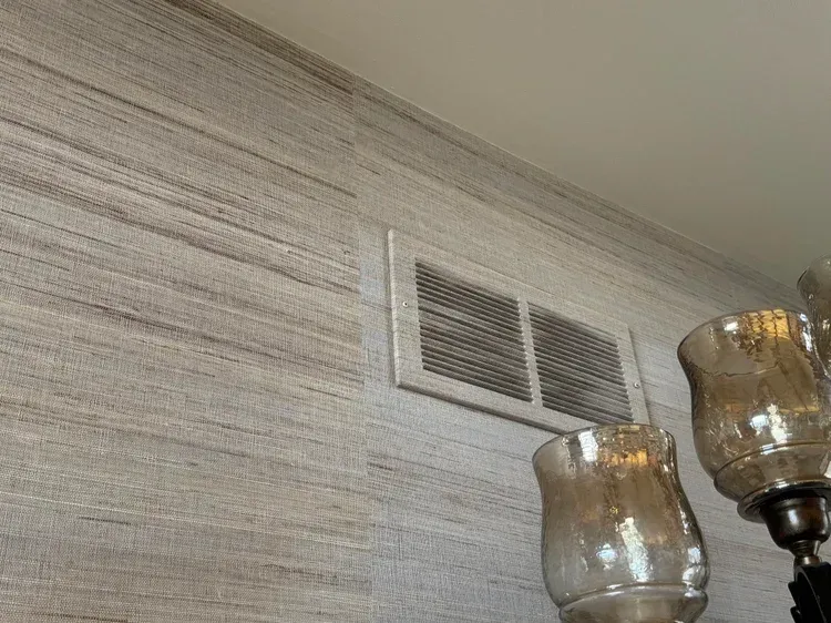 A tan textured wallpaper wall with an HVAC vent near the ceiling and a blurred glass lamp fixture in the foreground.