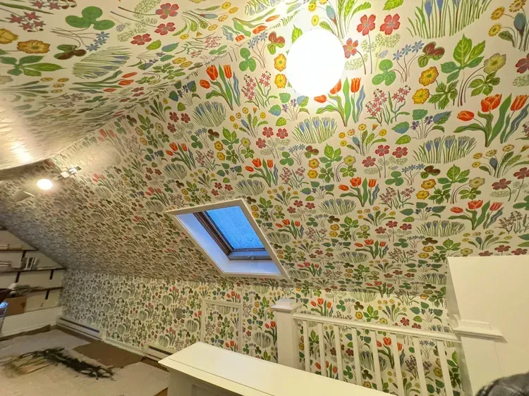 Attic room with slanted walls covered in colorful, busy floral wallpaper, a skylight, and white railing.