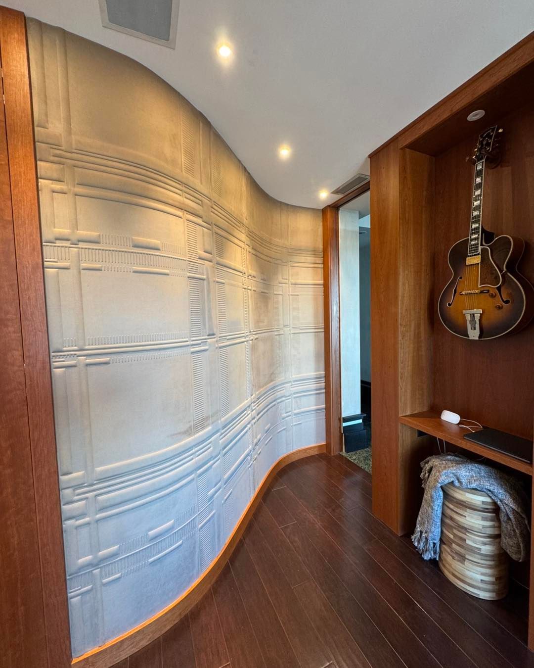 A curved, textured light wall beside a wood-paneled alcove displaying a sunburst-finish hollow-body guitar above a stool.