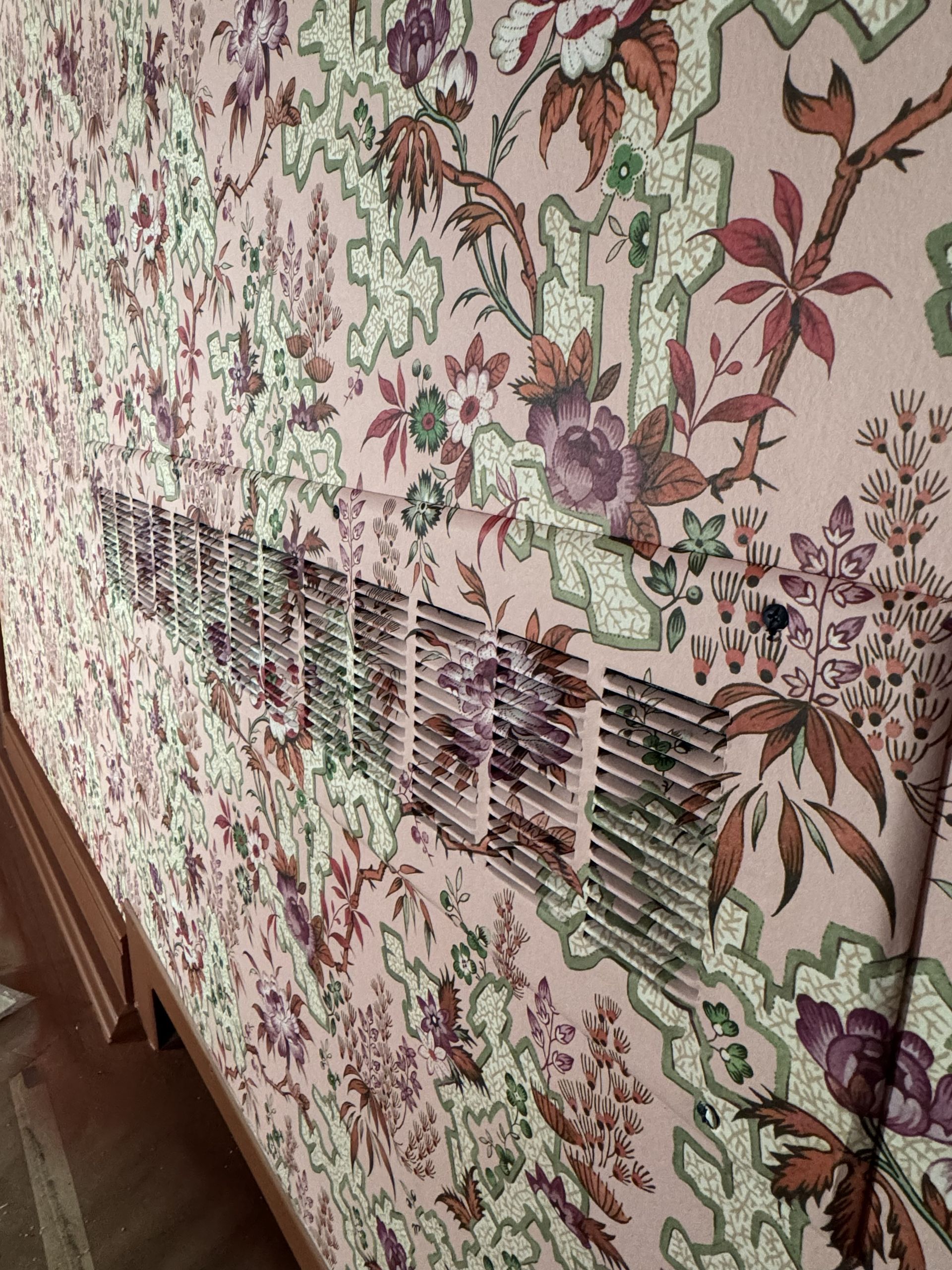 A horizontal metal wall vent grate is centered on a pale pink wall with intricate, vintage-style floral wallpaper.