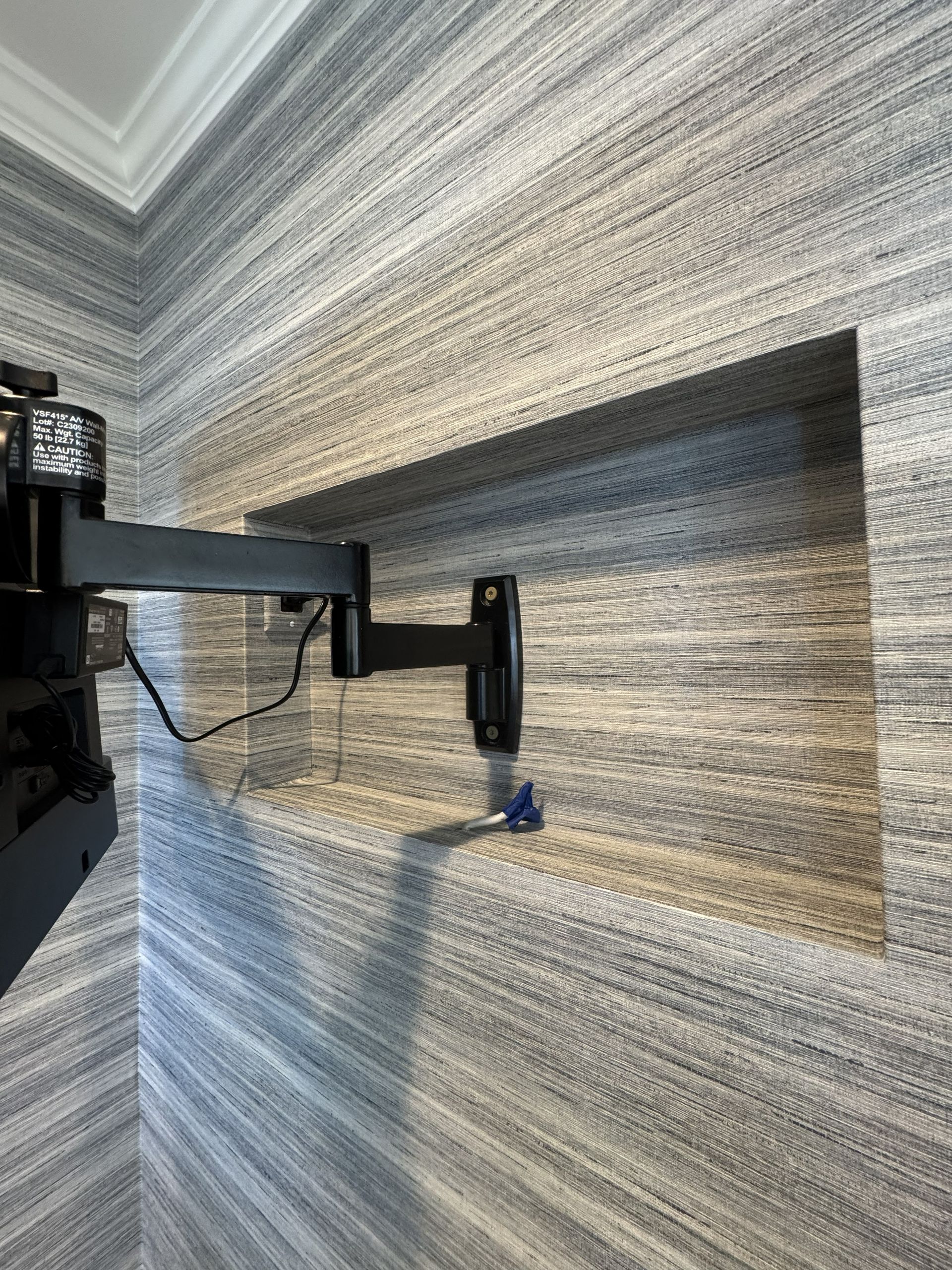 A black articulated TV wall mount attached inside a recessed wall niche with textured gray and brown horizontal wallpaper.
