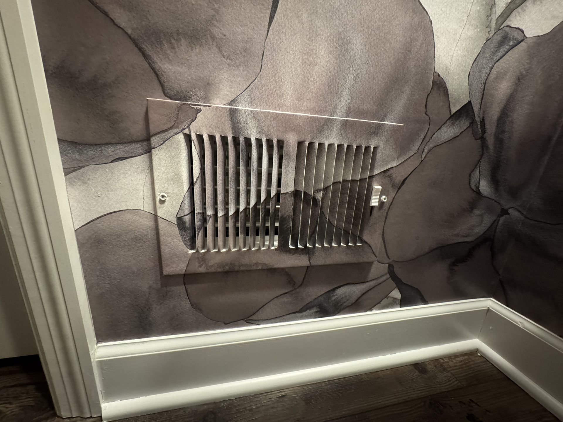 A metal HVAC floor register vent mounted on a wall with dark, floral-patterned wallpaper above white baseboards.