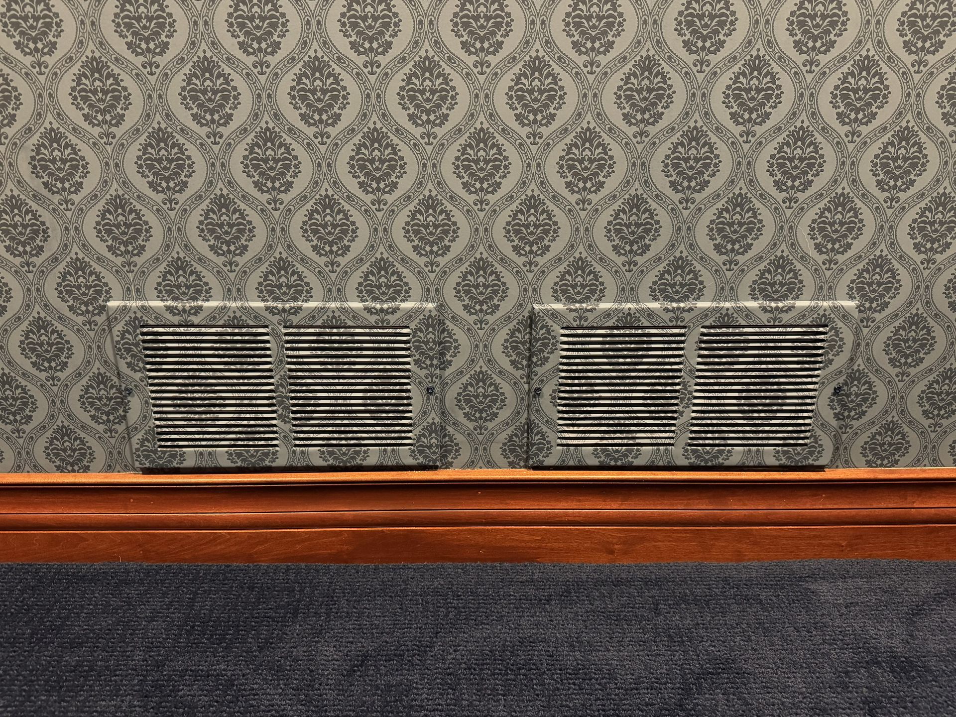 Two gray, rectangular air vents with horizontal slats, installed on a wall covered in gray patterned wallpaper.