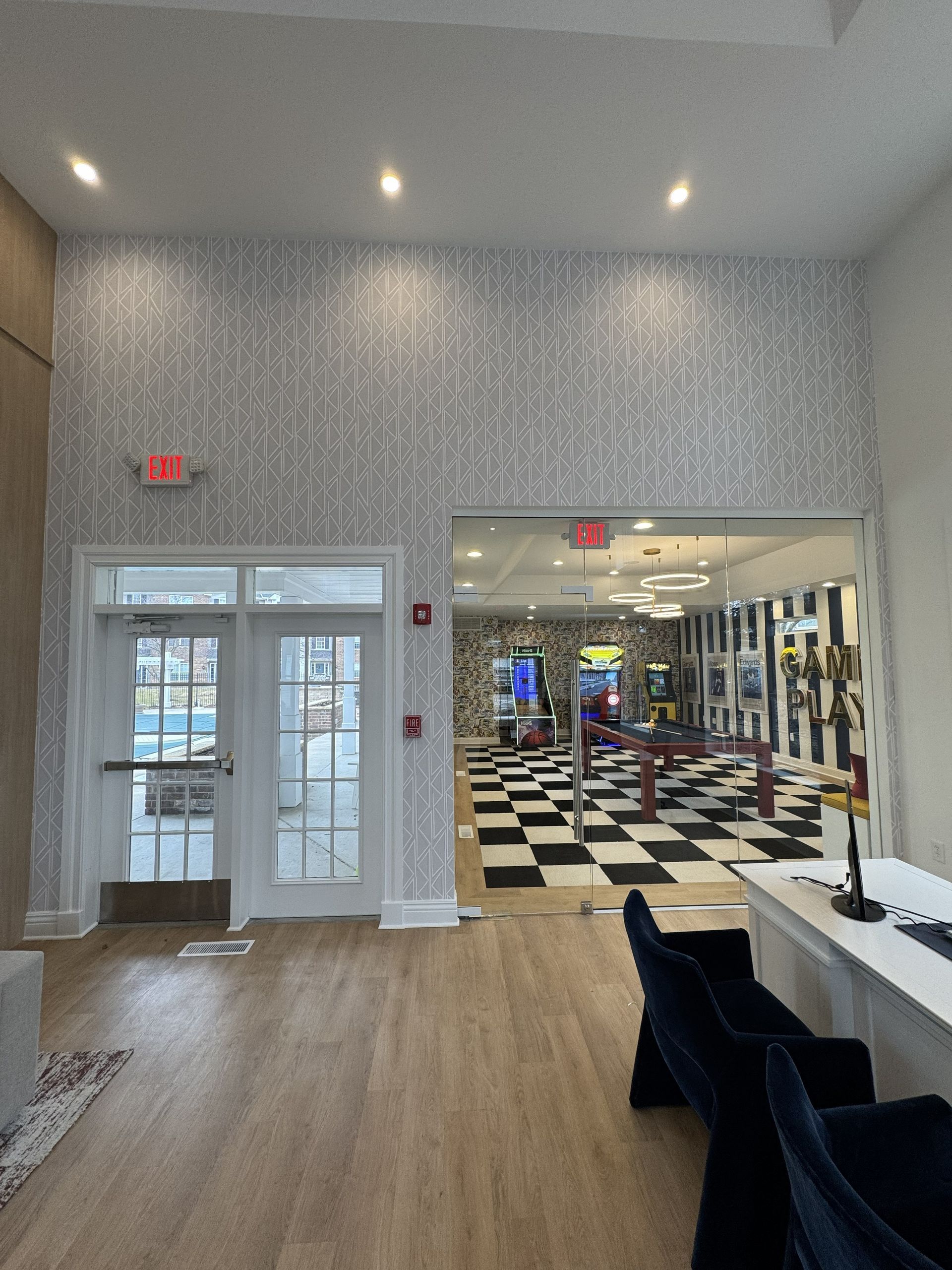 A lobby with light wood floors, a patterned wall, double glass doors to the outside, and a view into a game room.