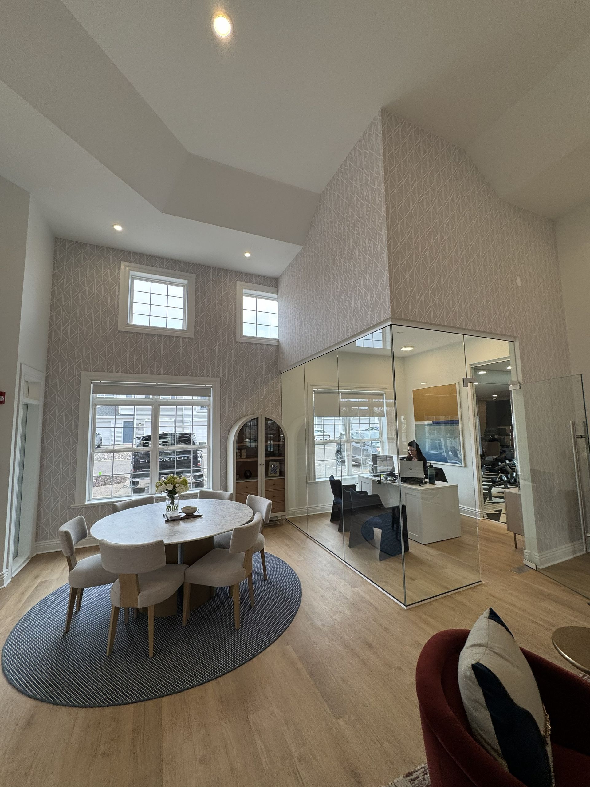 A bright dining room with a round table, blue patterned rug, and a large, mirrored wall reflecting an office space.