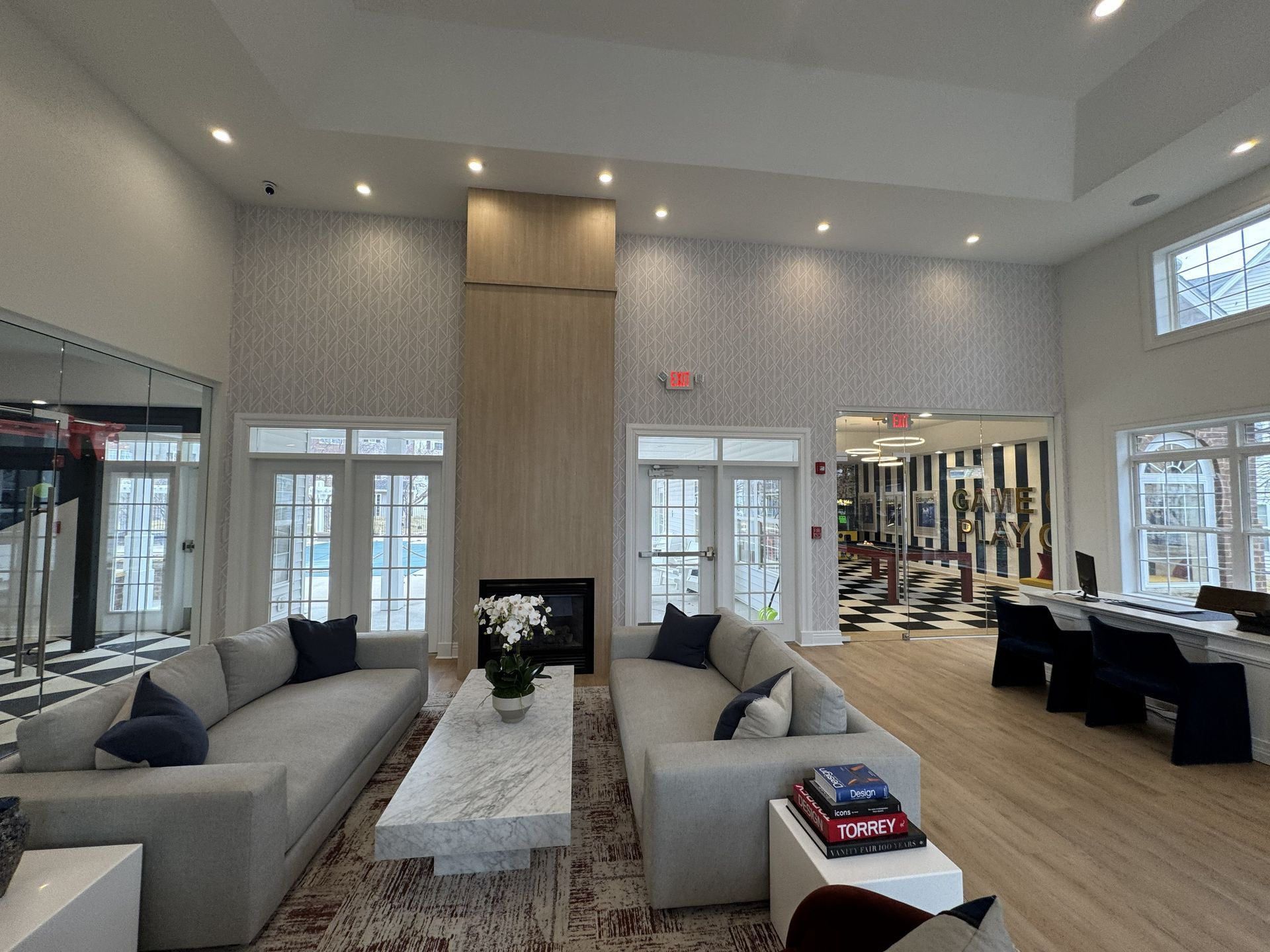A modern lobby with two grey sofas facing a fireplace, light wood paneling, marble coffee table, and wood-look flooring.