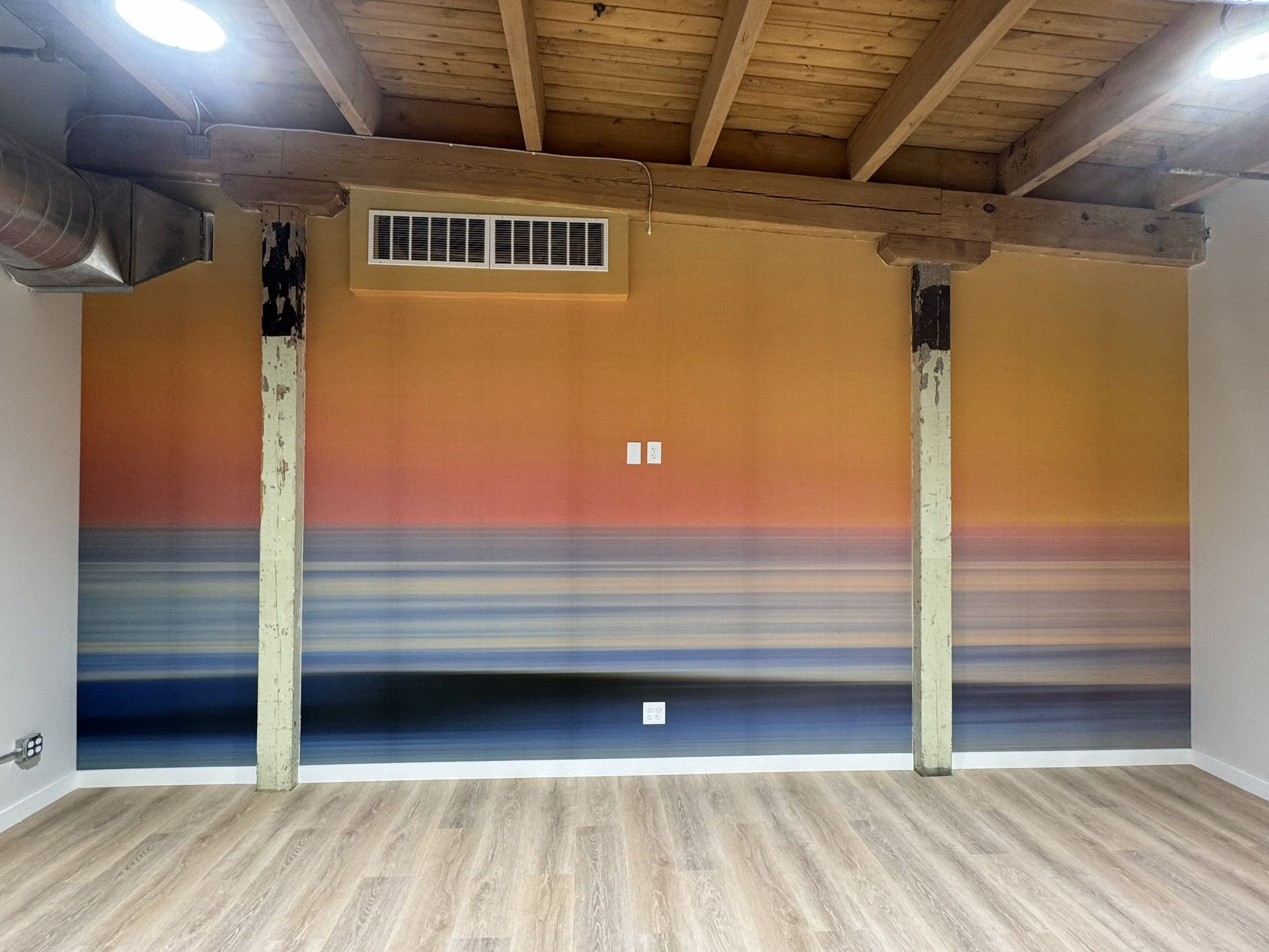 A sunset ocean mural on a wall between two wooden support pillars in a room with a wood-plank ceiling and floor.