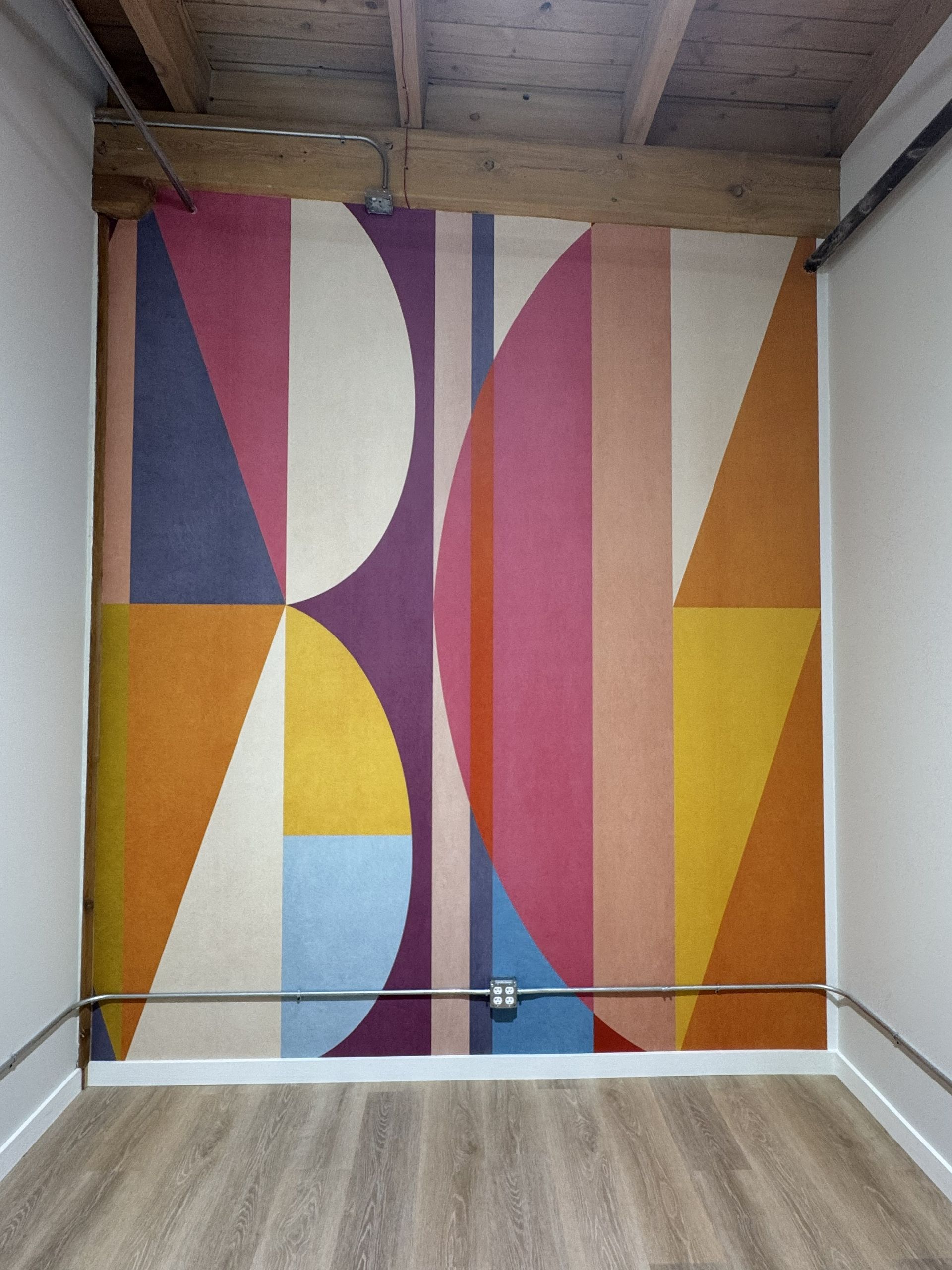 A colorful geometric mural on a wall featuring large semicircles and vertical stripes in muted pinks, yellows, and blues.