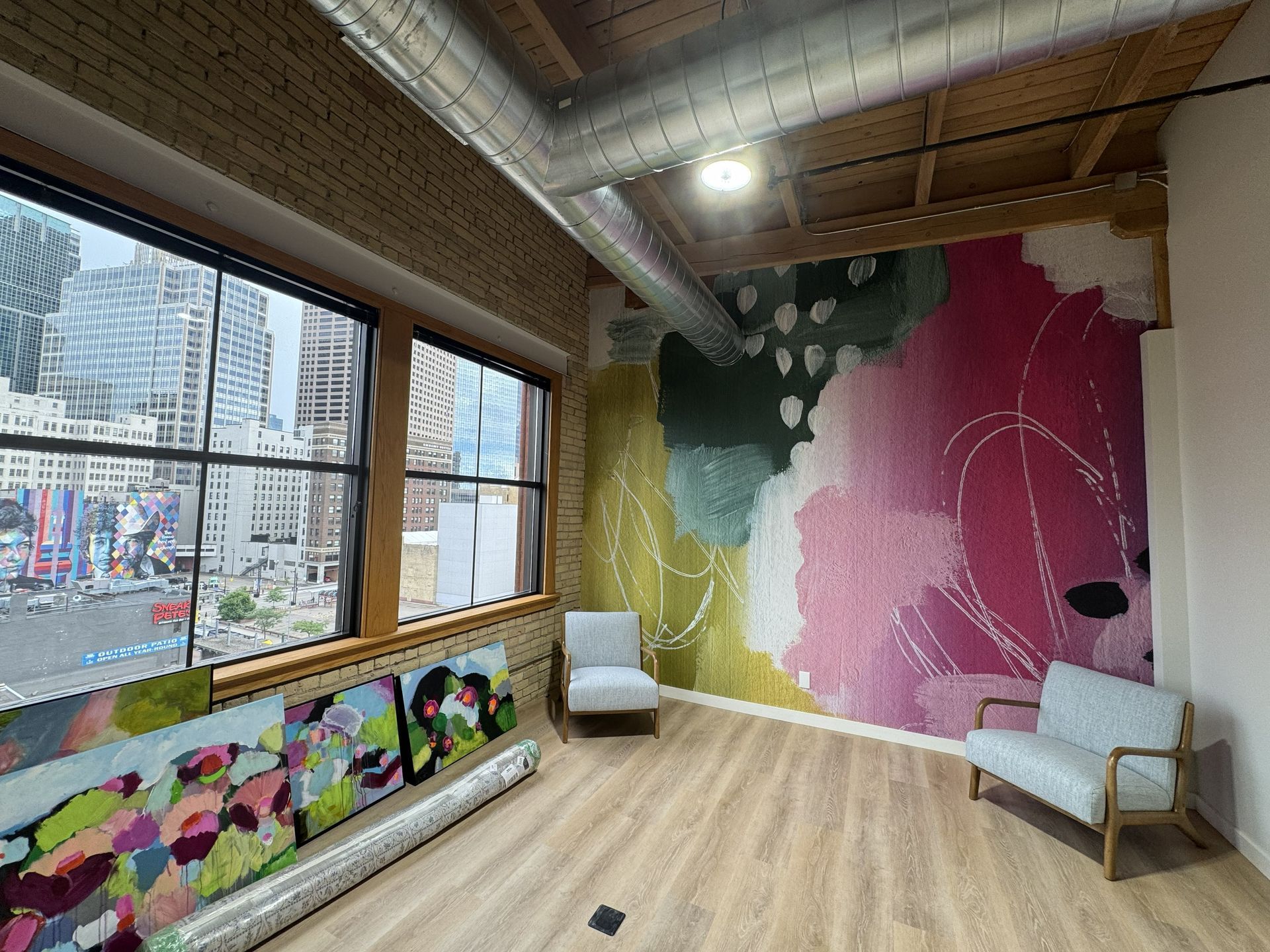 A bright, modern office corner with a colorful abstract mural, light wood floors, two chairs, and large city-view windows.