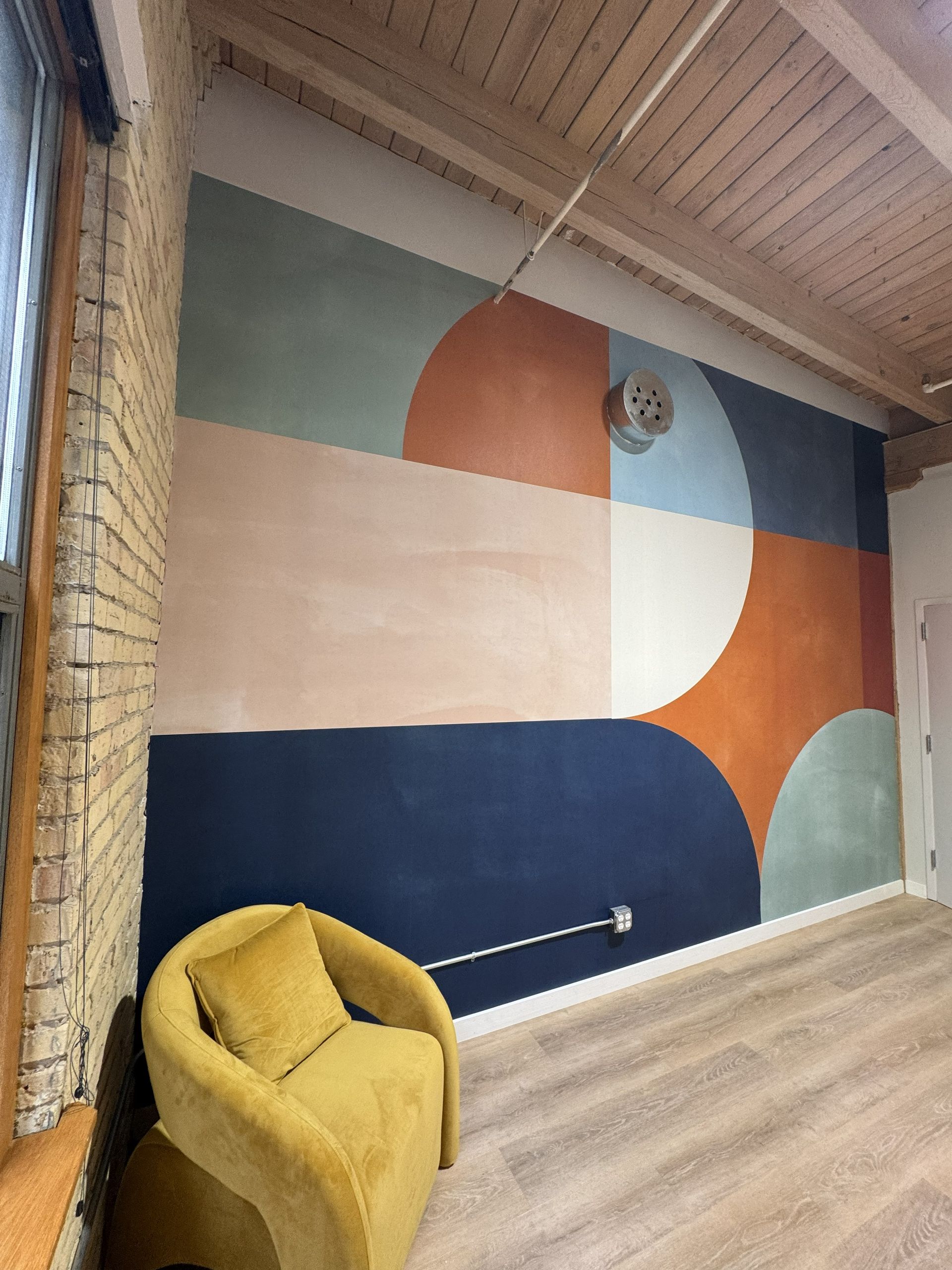 A room wall featuring a large geometric mural in muted blue, orange, cream, and green with a yellow chair in front.