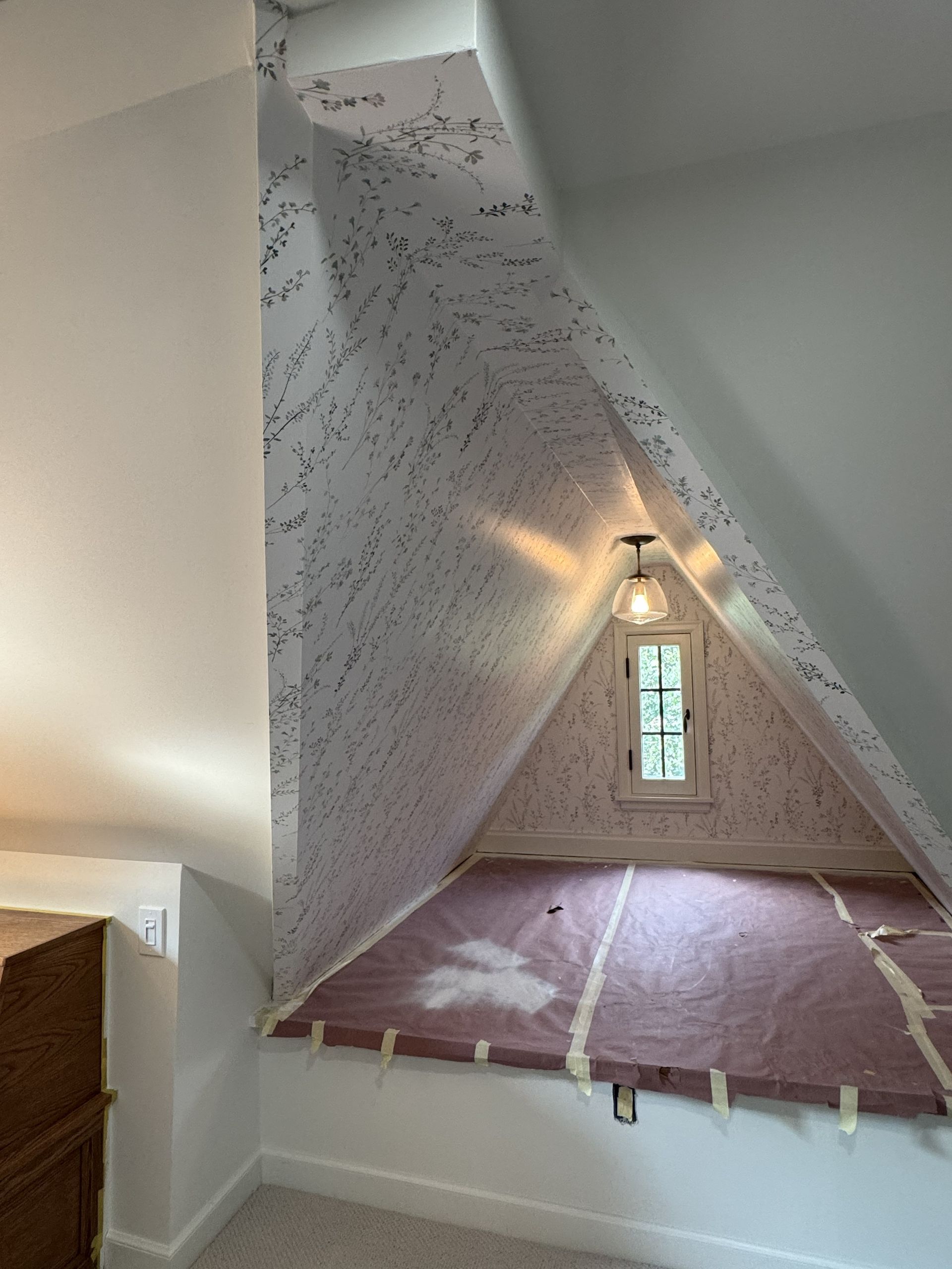A triangular window nook with patterned wallpaper, a pendant light, and a boarded-up platform bed in an attic space.