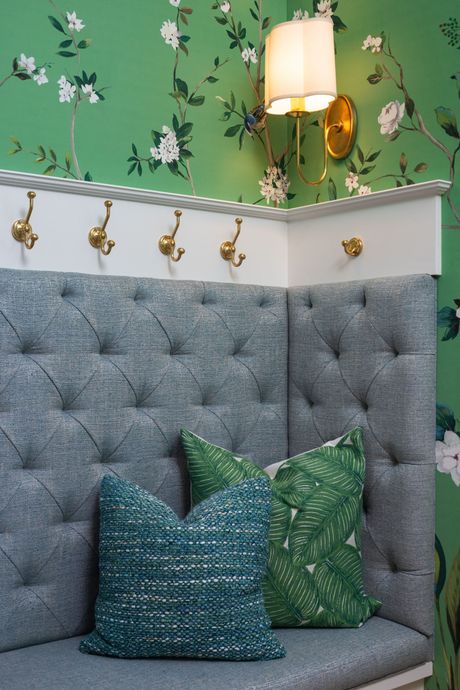 A corner upholstered bench with button tufting, green floral wallpaper, gold coat hooks, and decorative throw pillows.