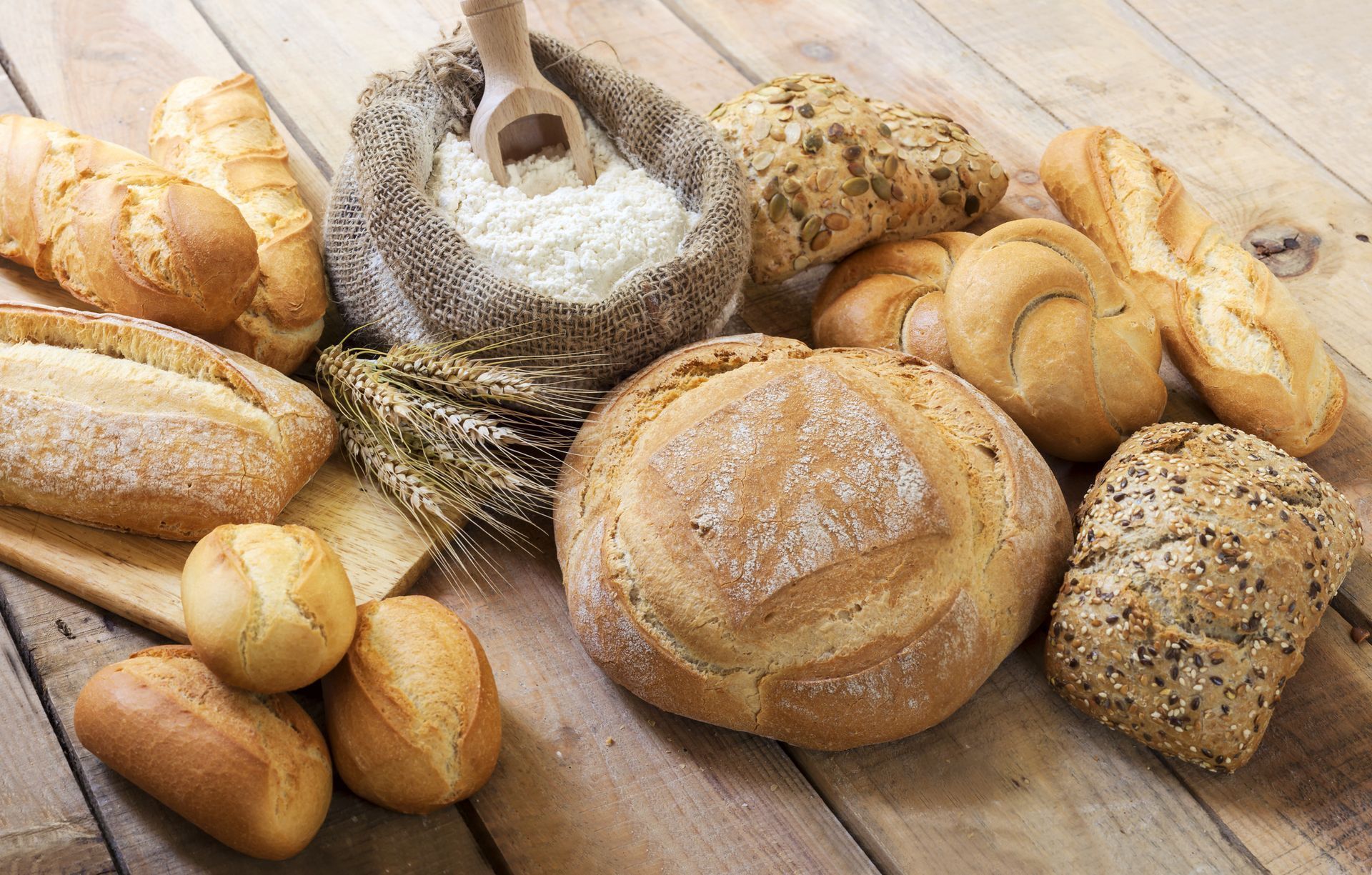 Su un tavolo di legno si trovano diversi tipi di pane e panini rustici, tra cui una pagnotta infarinata in un cestino.