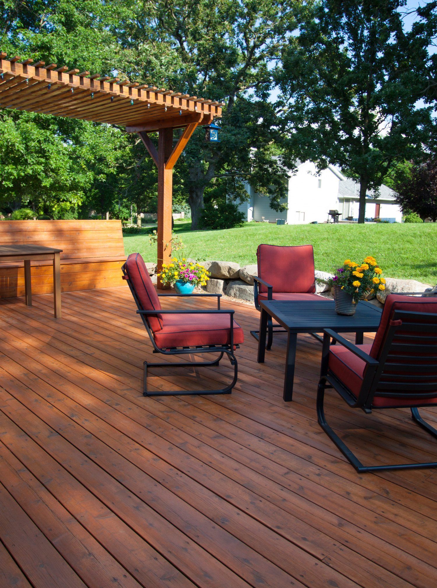 A wooden backyard deck with a pergola, a bench, a table, and red chairs surrounded by green trees and lawn.