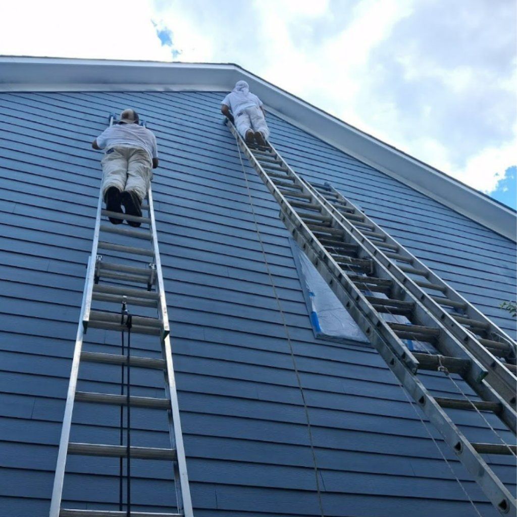 Two workers in white suits climb tall ladders against a blue-sided house exterior.