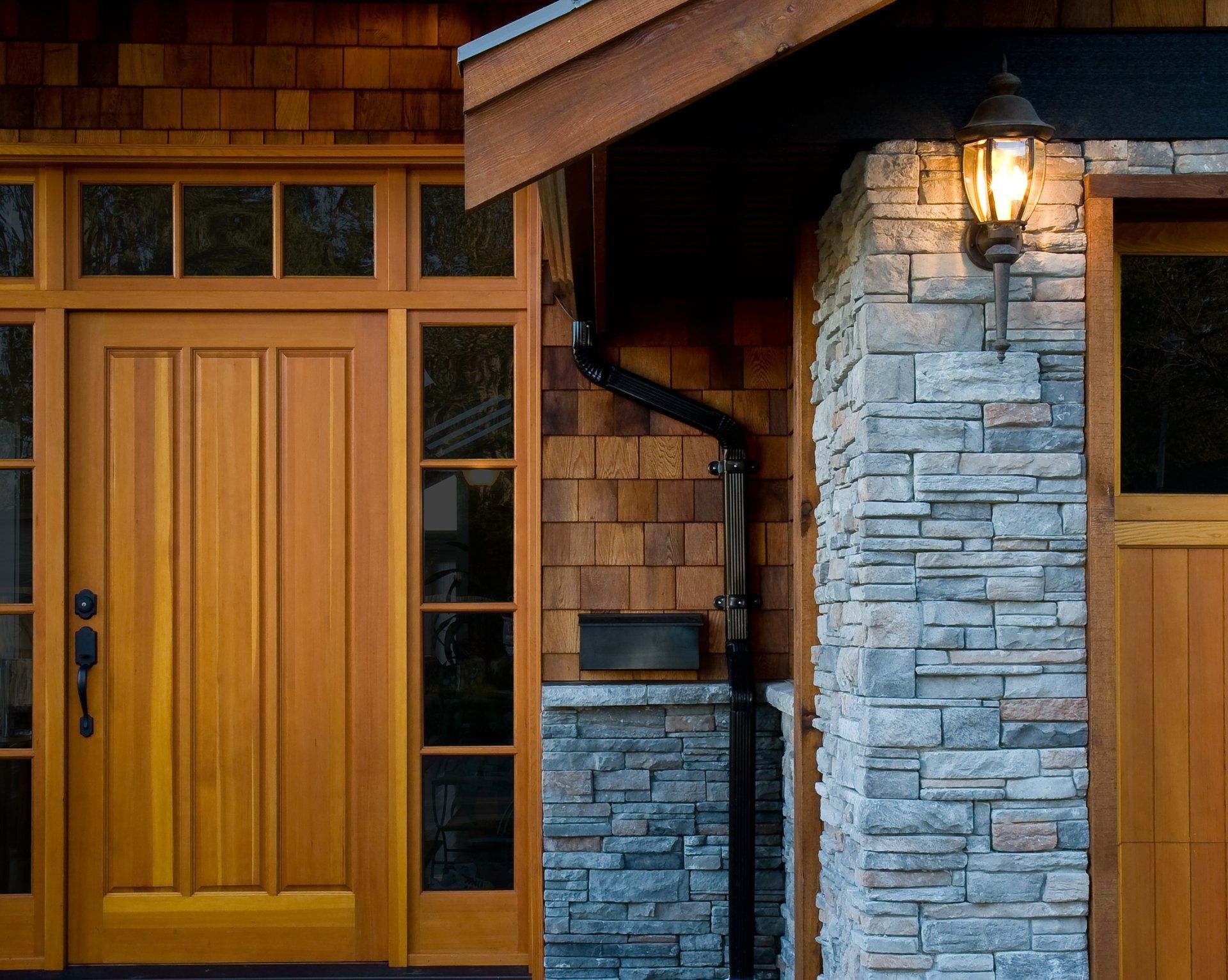 A front entrance with a wooden door, sidelights, stone pillar, and cedar shingles, illuminated by an exterior lamp.