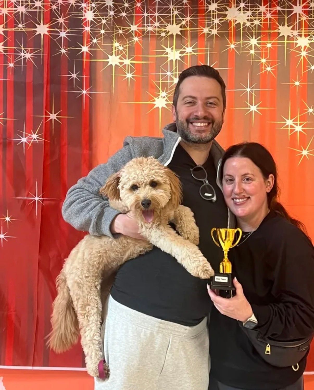 Smiling couple holding a fluffy dog and a trophy before a red, sparkly backdrop.