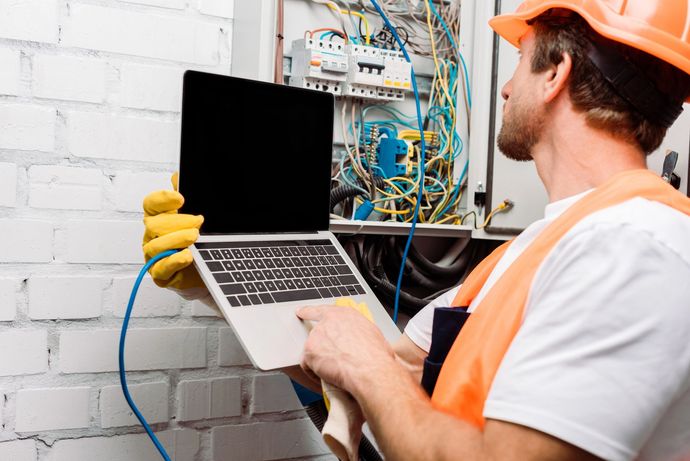Una persona con elmetto e giubbotto ad alta visibilità utilizza un computer portatile per effettuare la manutenzione di un quadro elettrico contro un muro di mattoni bianchi.