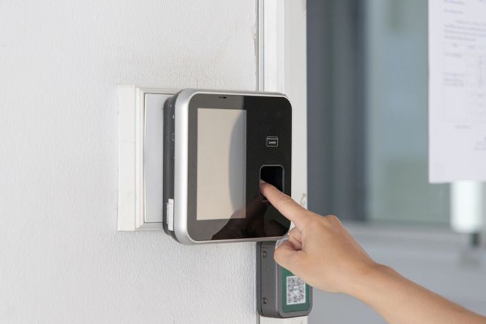 Una mano appoggia un dito su uno scanner biometrico per impronte digitali montato su una parete bianca.