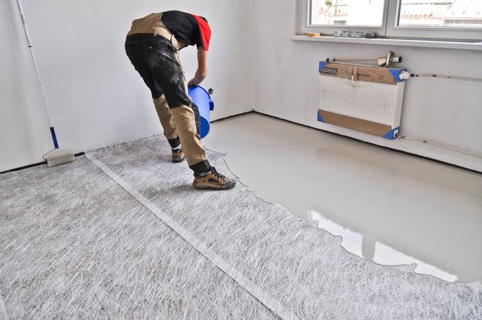 Un operaio versa del composto autolivellante da un secchio blu su un pavimento grigio in una stanza in fase di ristrutturazione.