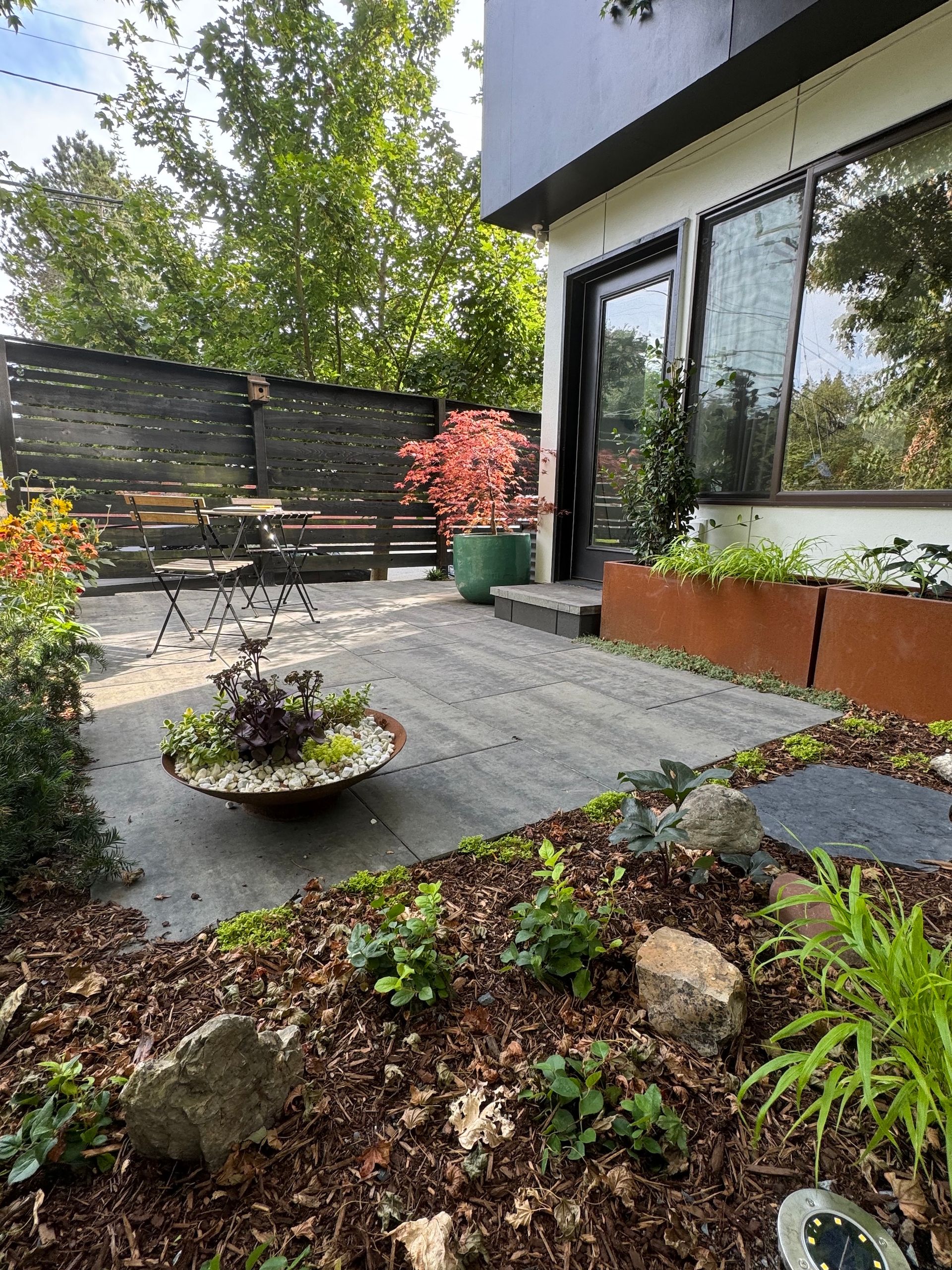 Modern patio with gray pavers, potted plants, bench seating, and glass doors beside a landscaped garden.