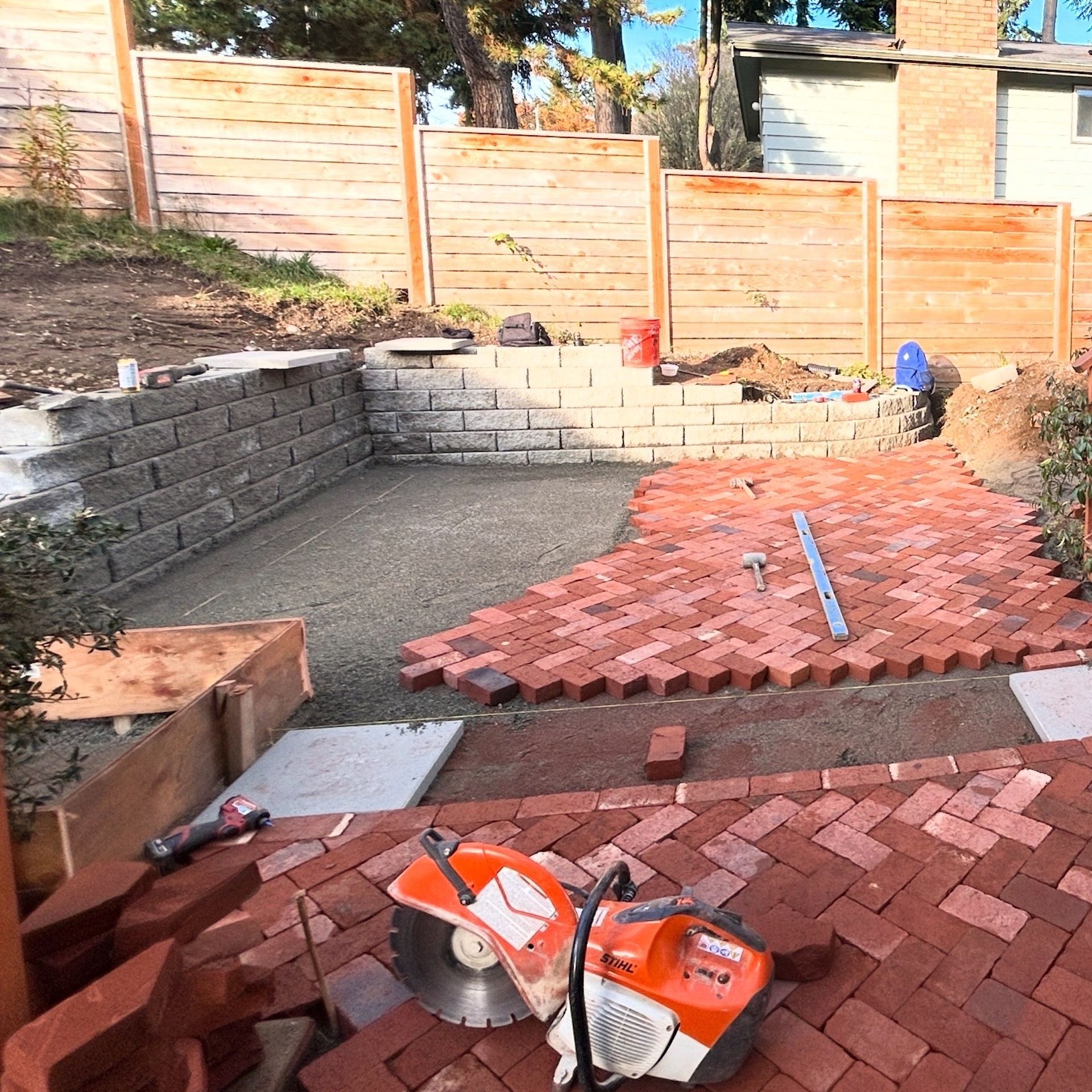 Backyard patio under construction with stacked red pavers, retaining wall, and tools on the ground
