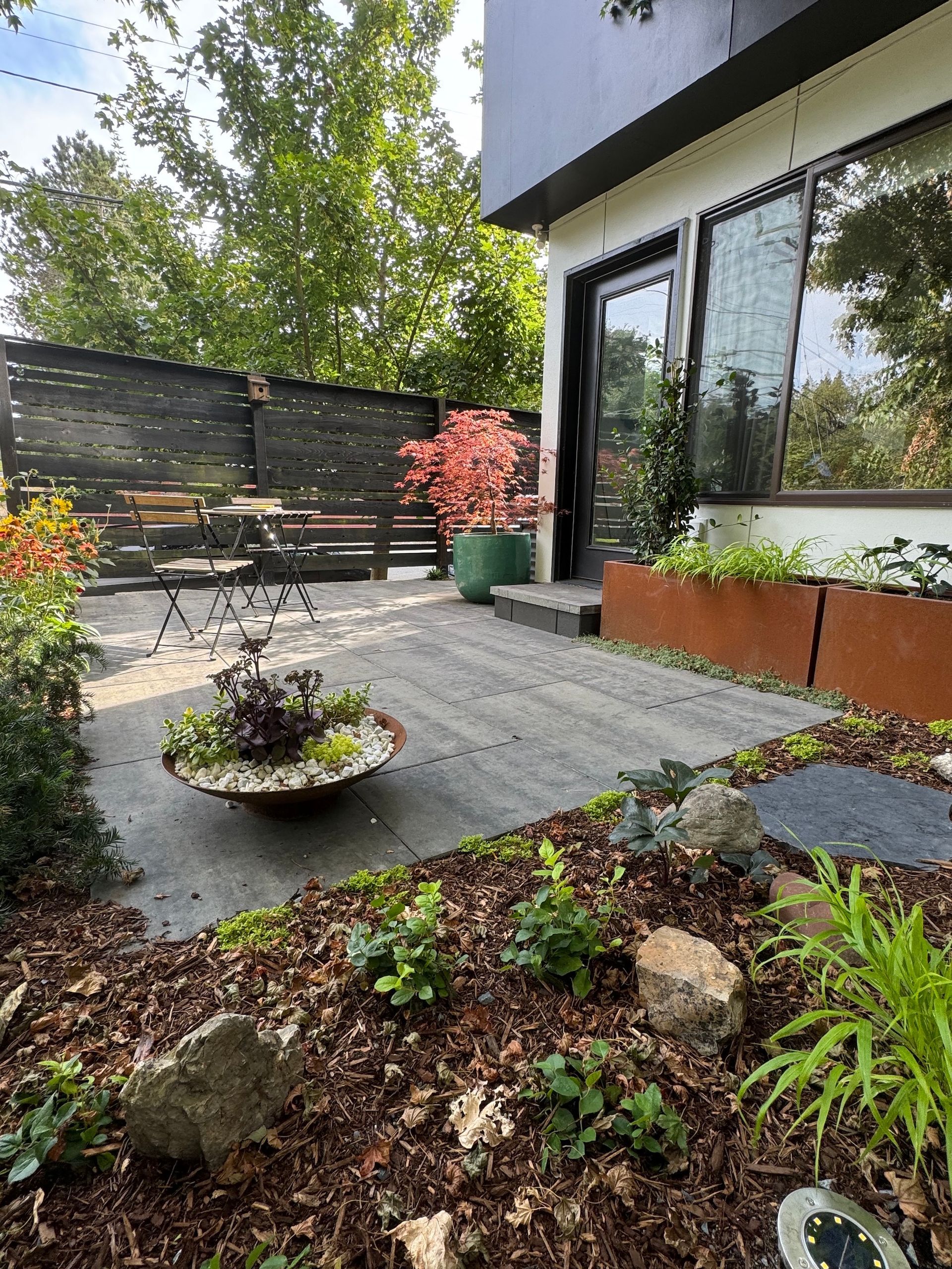 Modern patio with stone pavers, outdoor seating, potted plants, and glass doors beside a garden bed