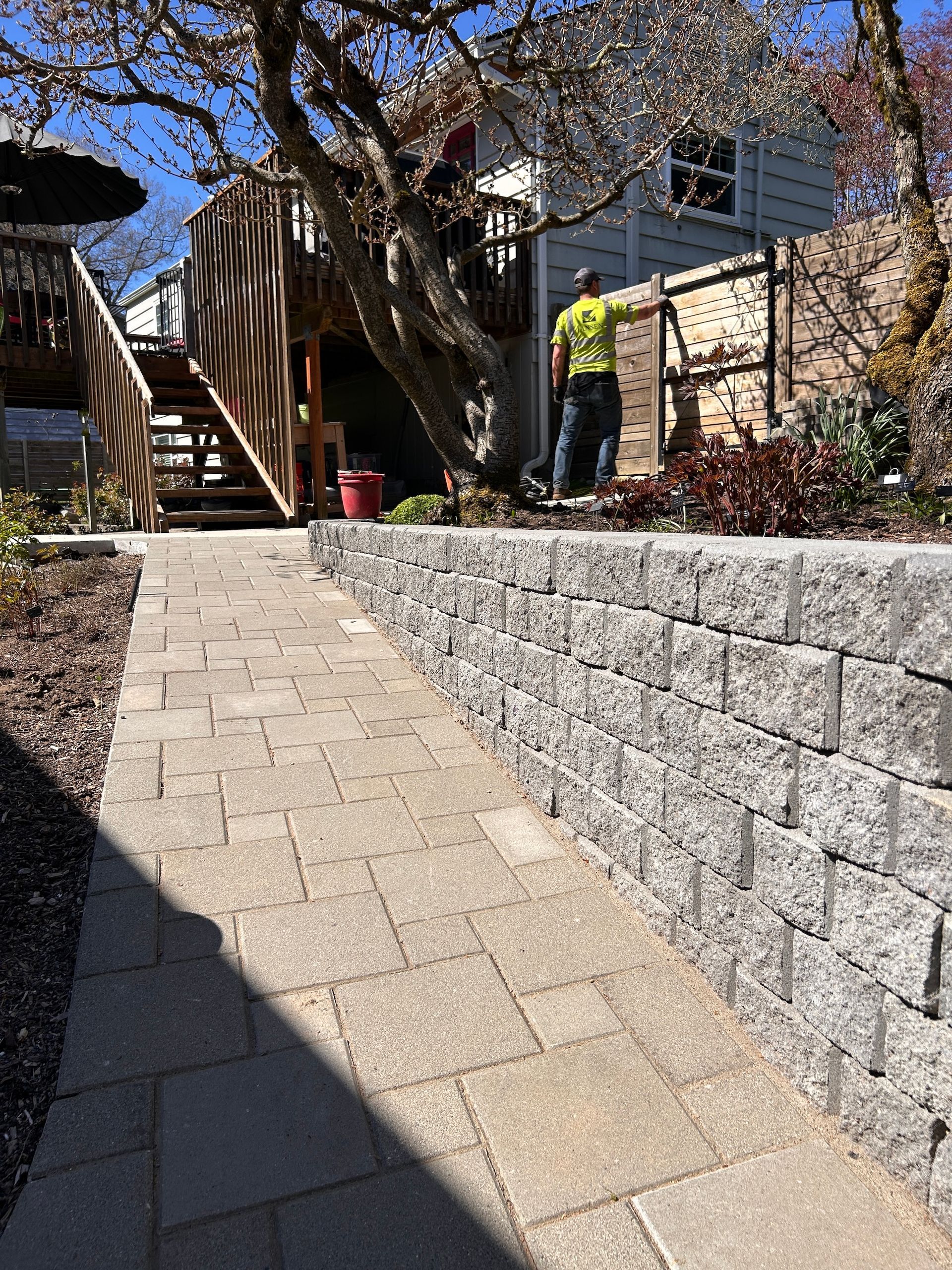 Stone walkway beside a gray retaining wall, leading past stairs and a person in a yellow jacket in a sunny yard