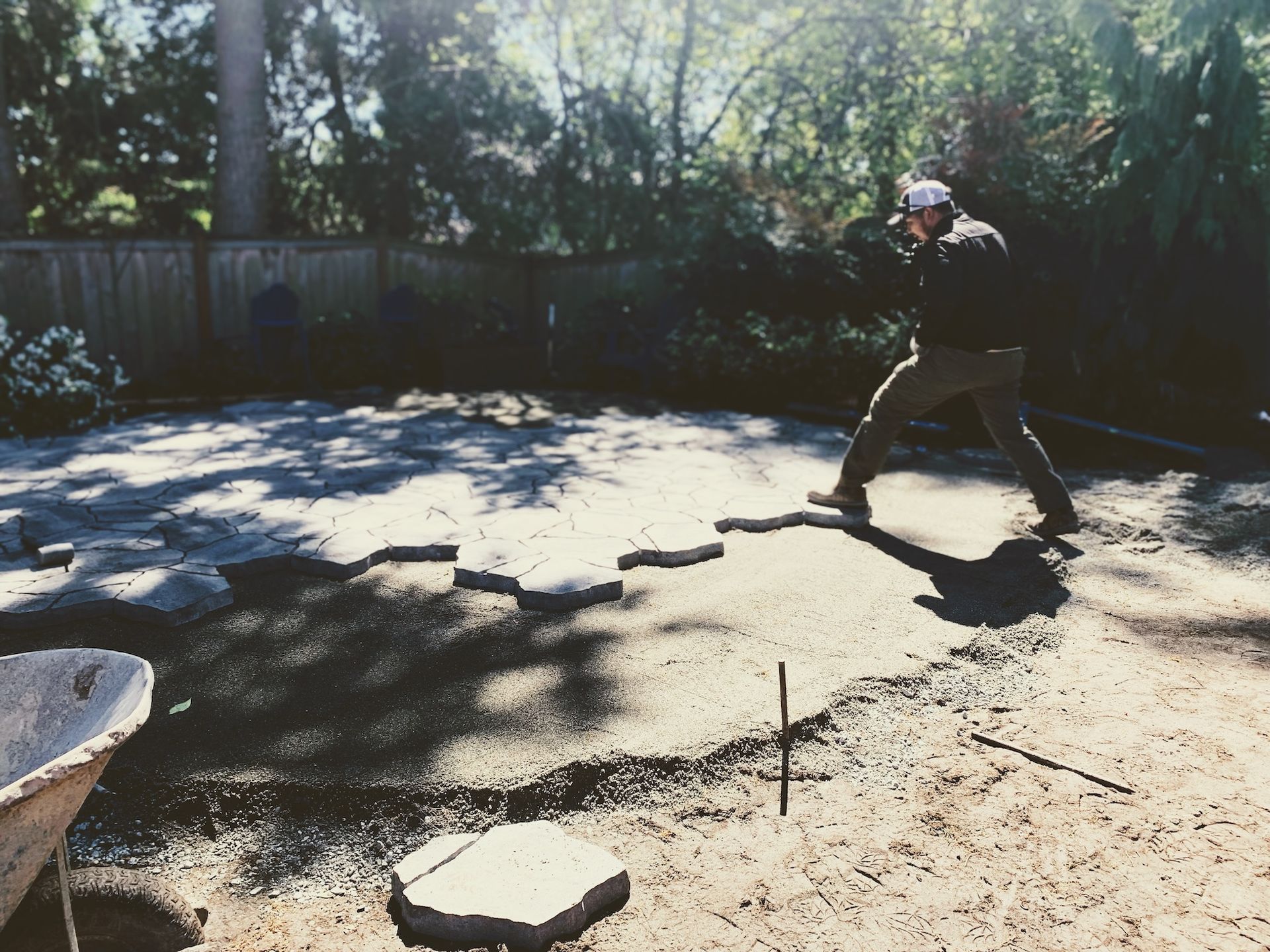 Person in dark clothing standing in a sunny backyard, aiming a paintball gun near stepping stones.