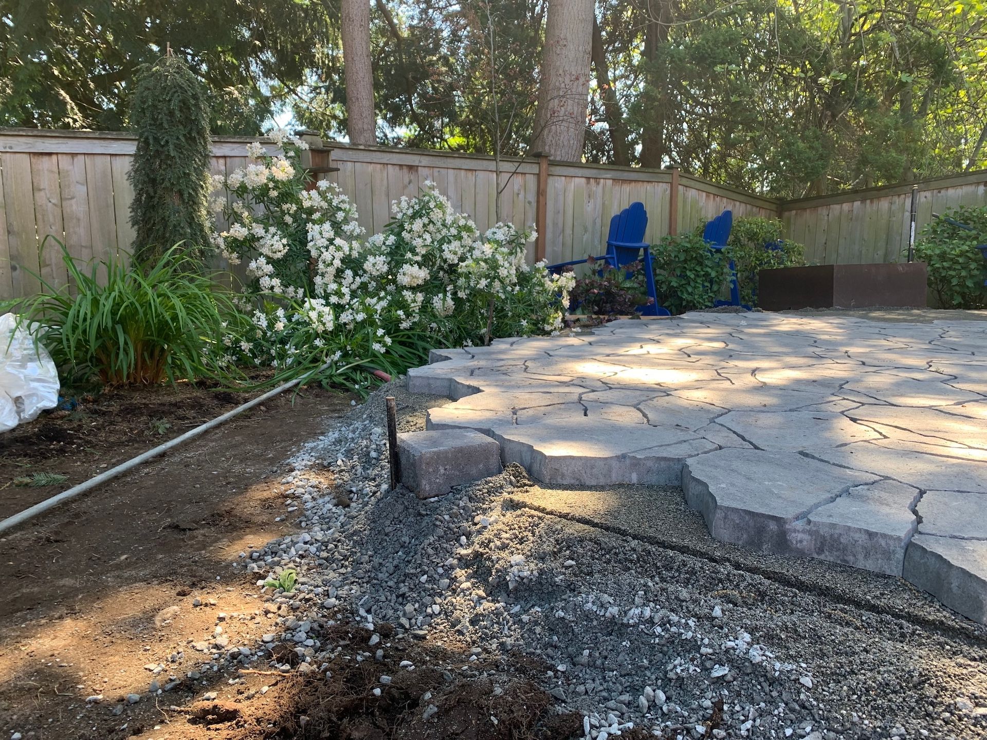 Cracked stone patio beside a garden bed and wooden fence, with white flowers and blue chairs in the background