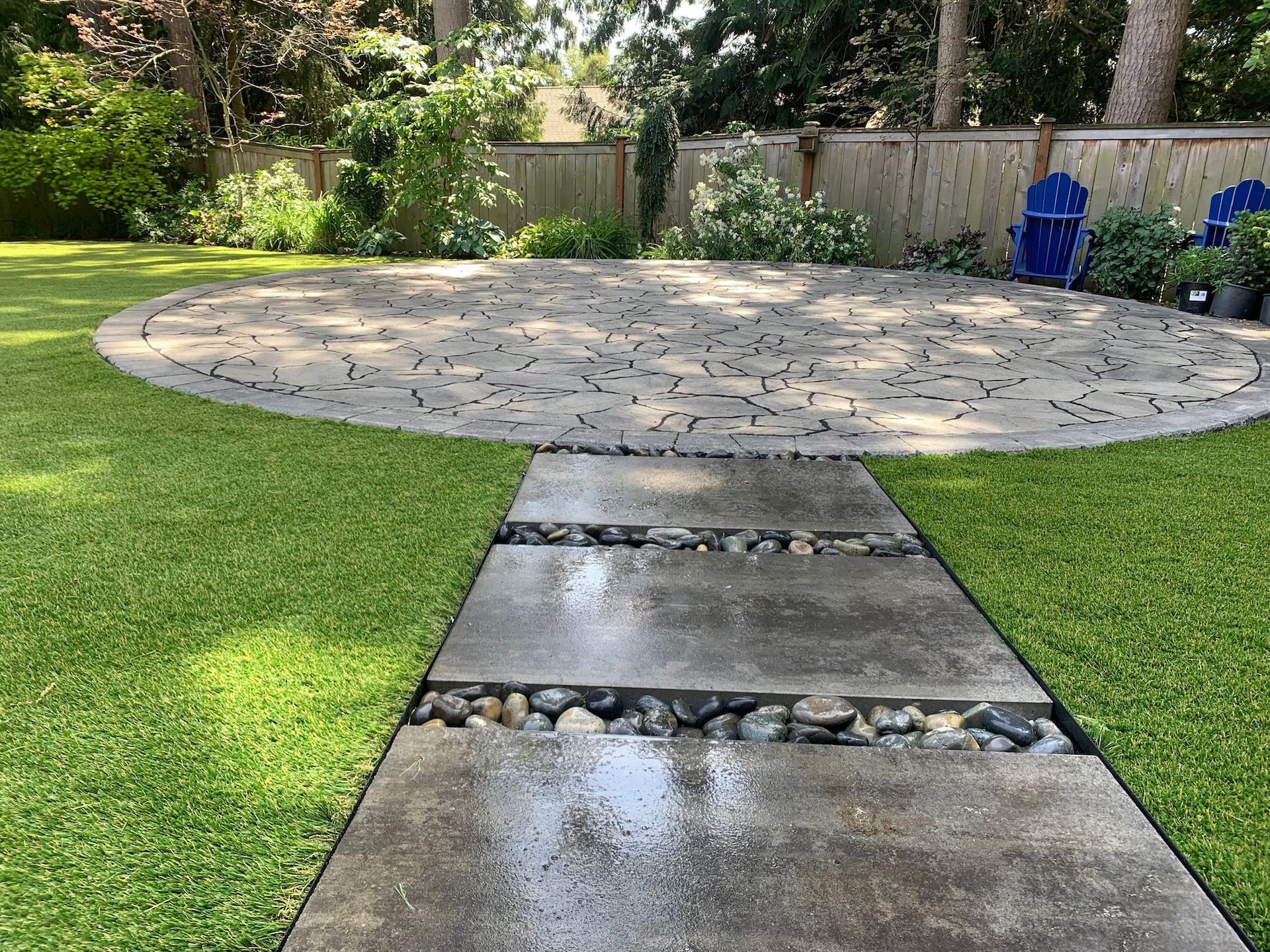 Backyard patio with a circular stone seating area and wet concrete path through green lawn