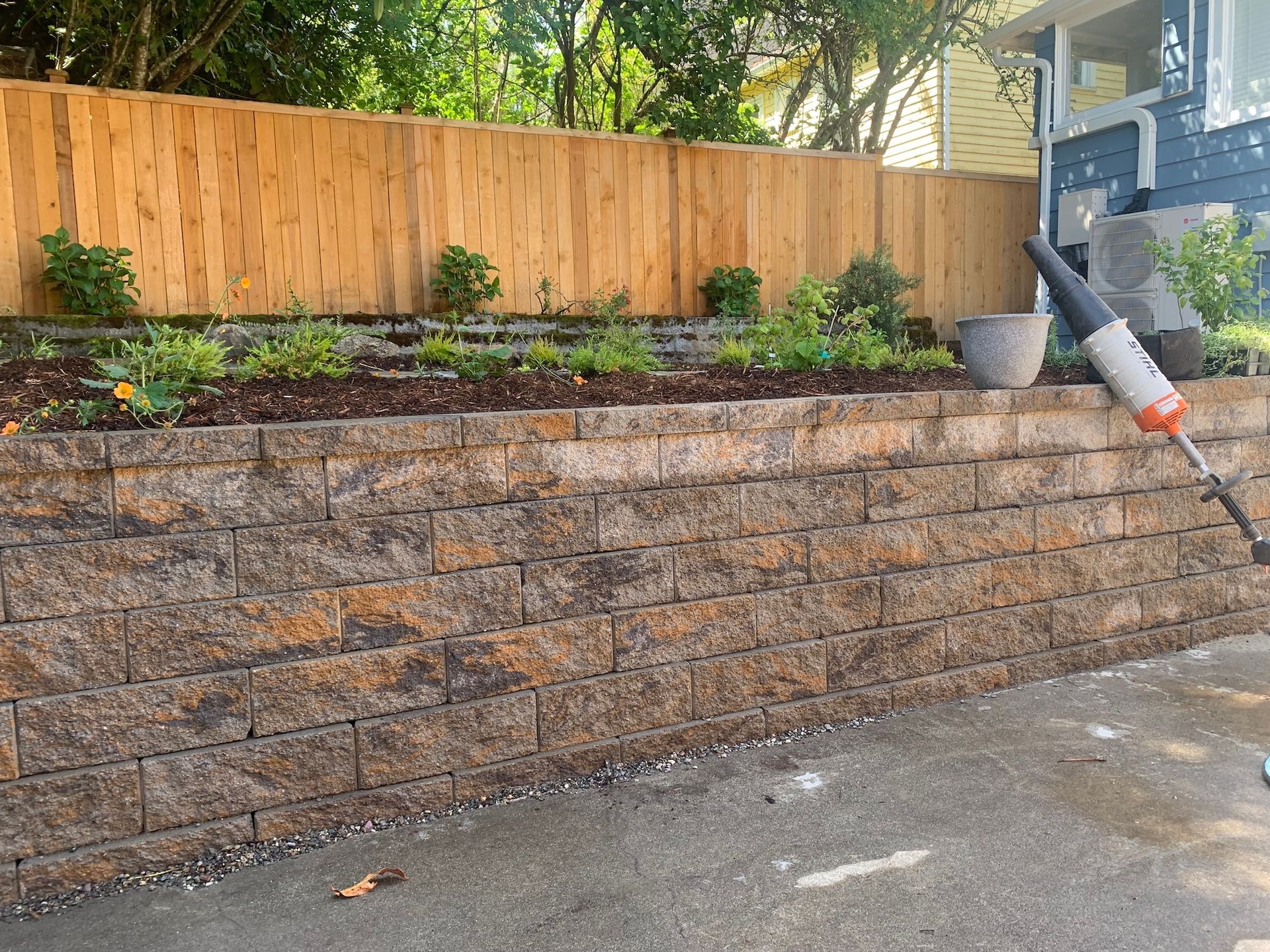 Stone retaining wall with garden beds and wooden fence above, beside a wet driveway and house exterior