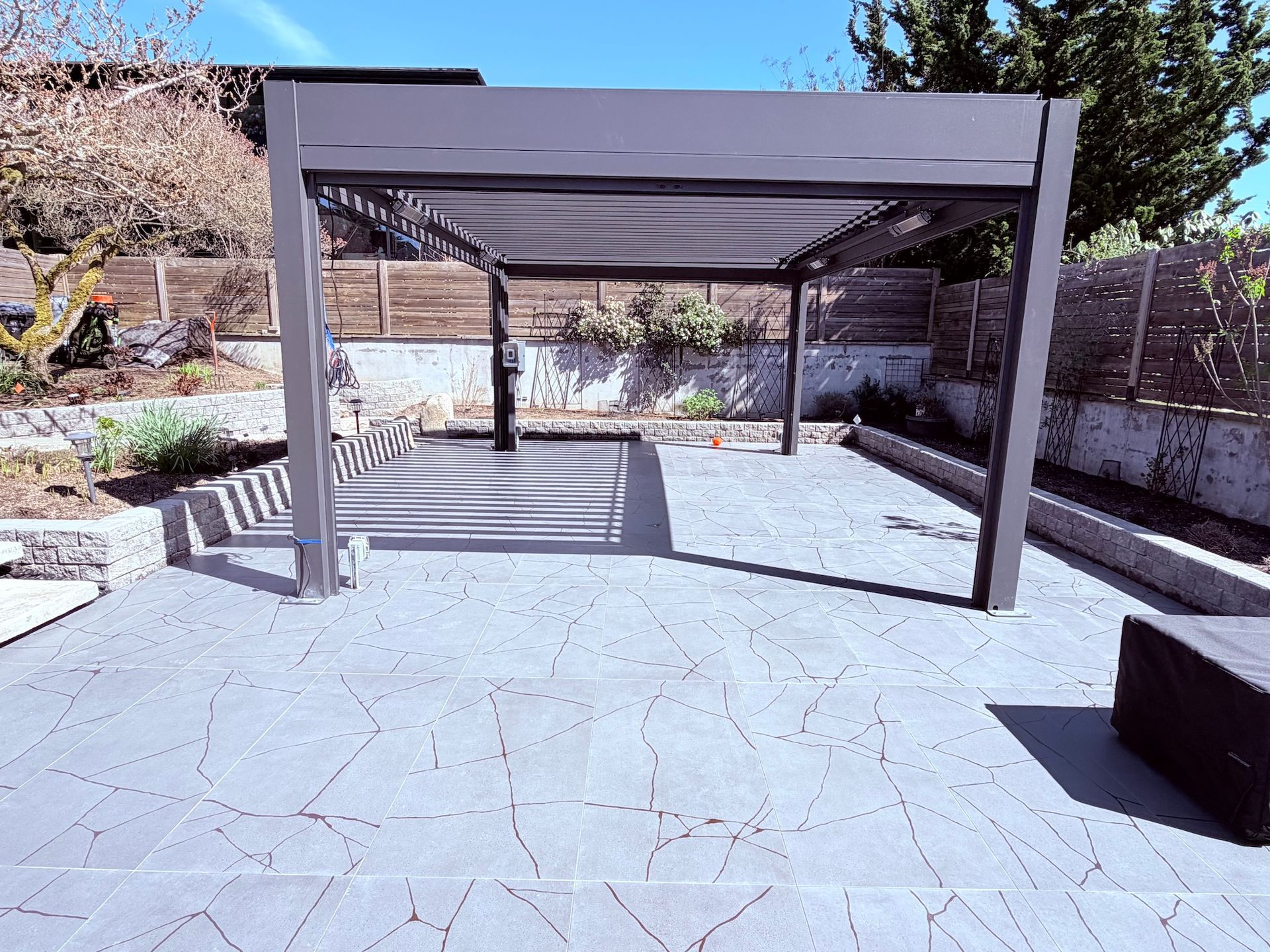 Outdoor patio with a gray pergola, stone paving, and benches in a sunny landscaped courtyard