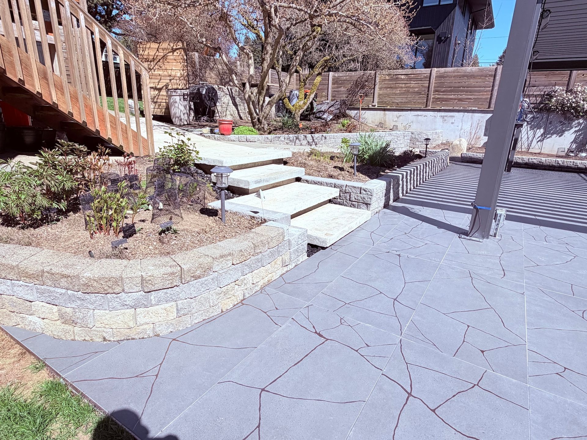 Stone patio and steps in a sunlit garden beside a wooden deck and house.