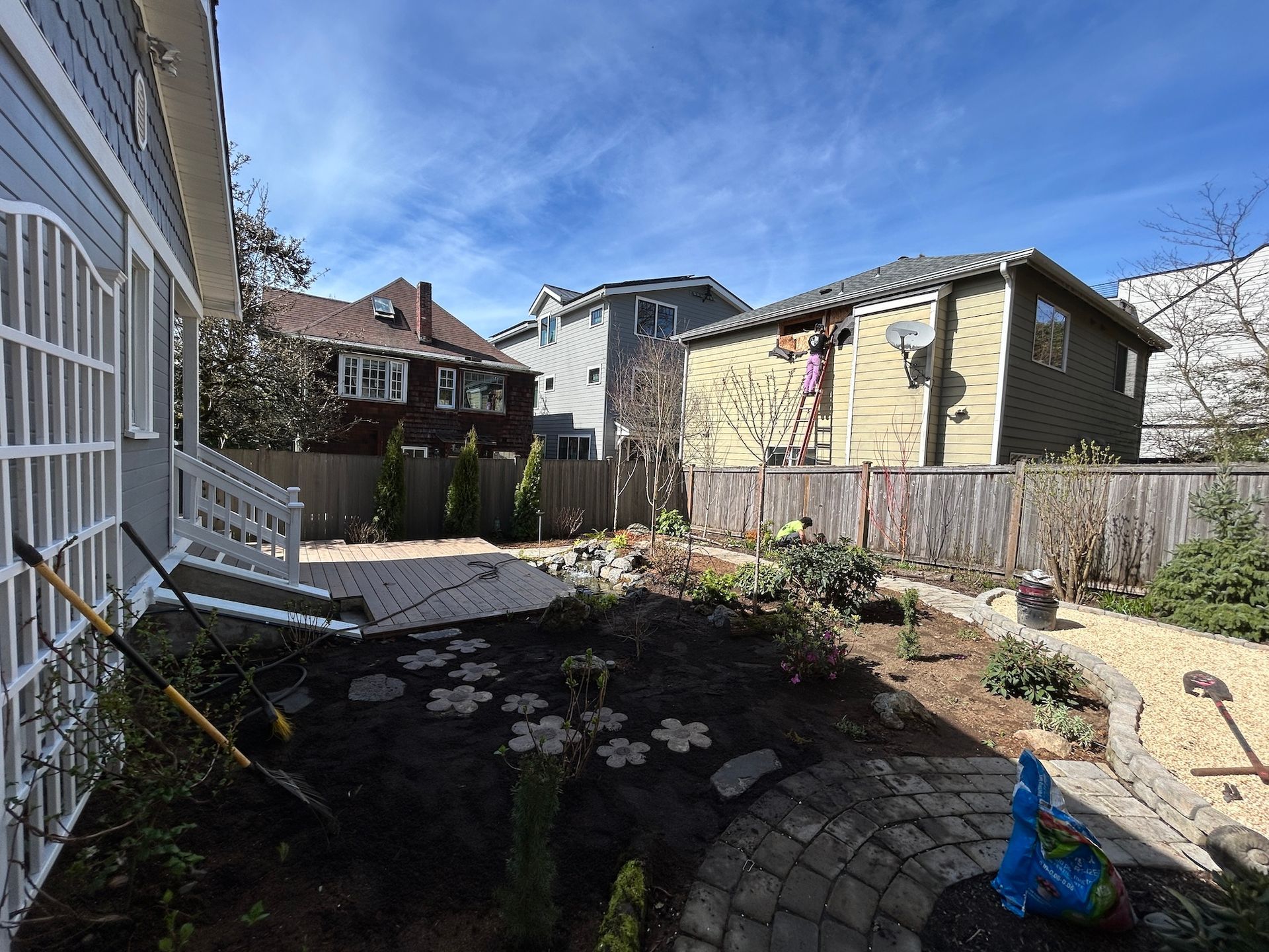 Backyard garden with raised beds, a patio, wooden fence, and neighboring houses under a blue sky
