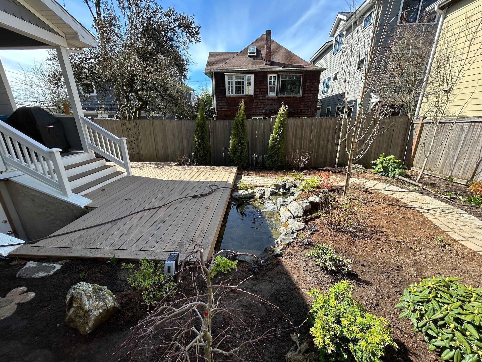 Backyard garden with deck, small pond, rocks, shrubs, and a house in the background