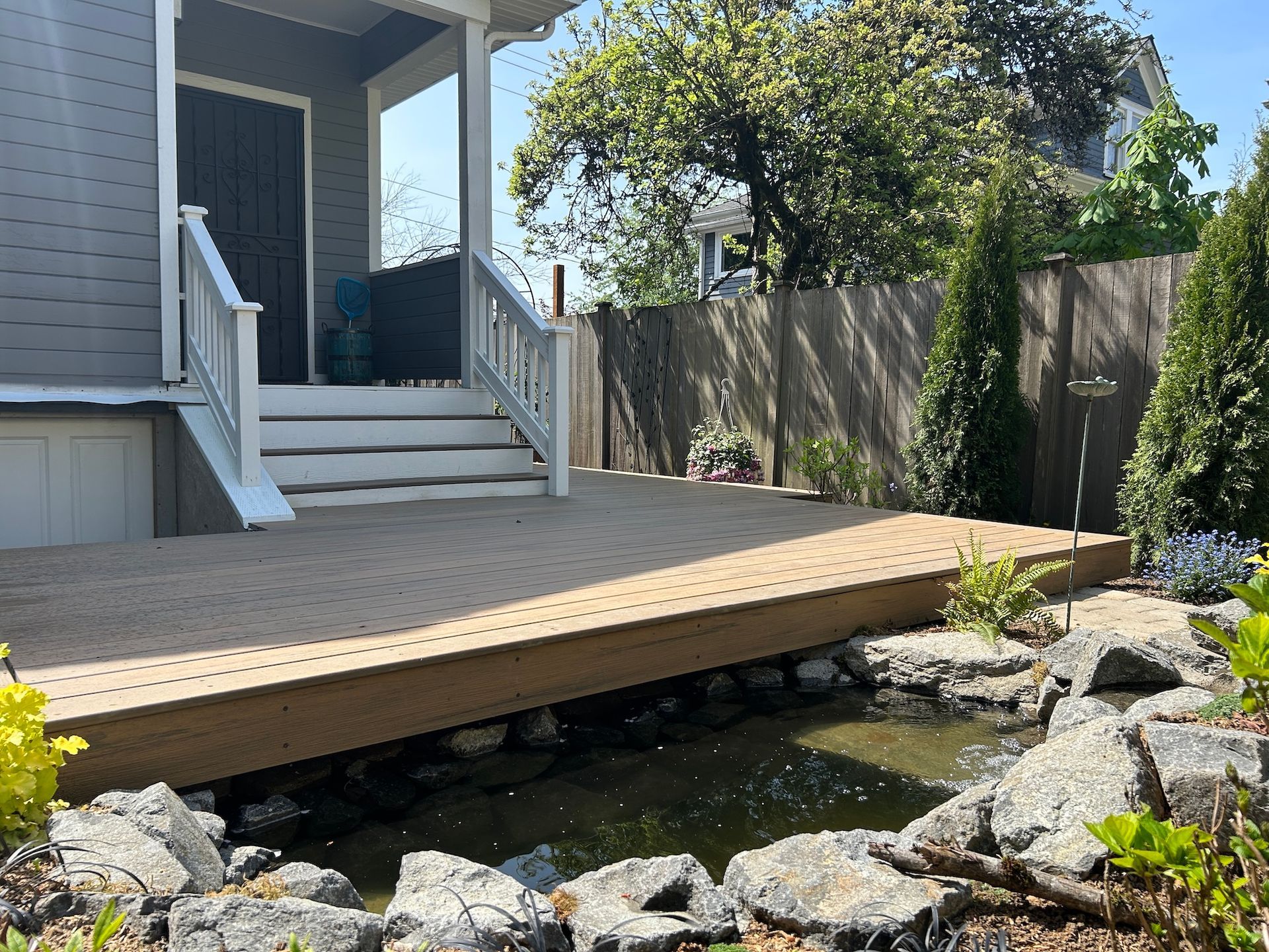 House deck with stairs overlooking a small rocky pond and garden fence in bright sunlight