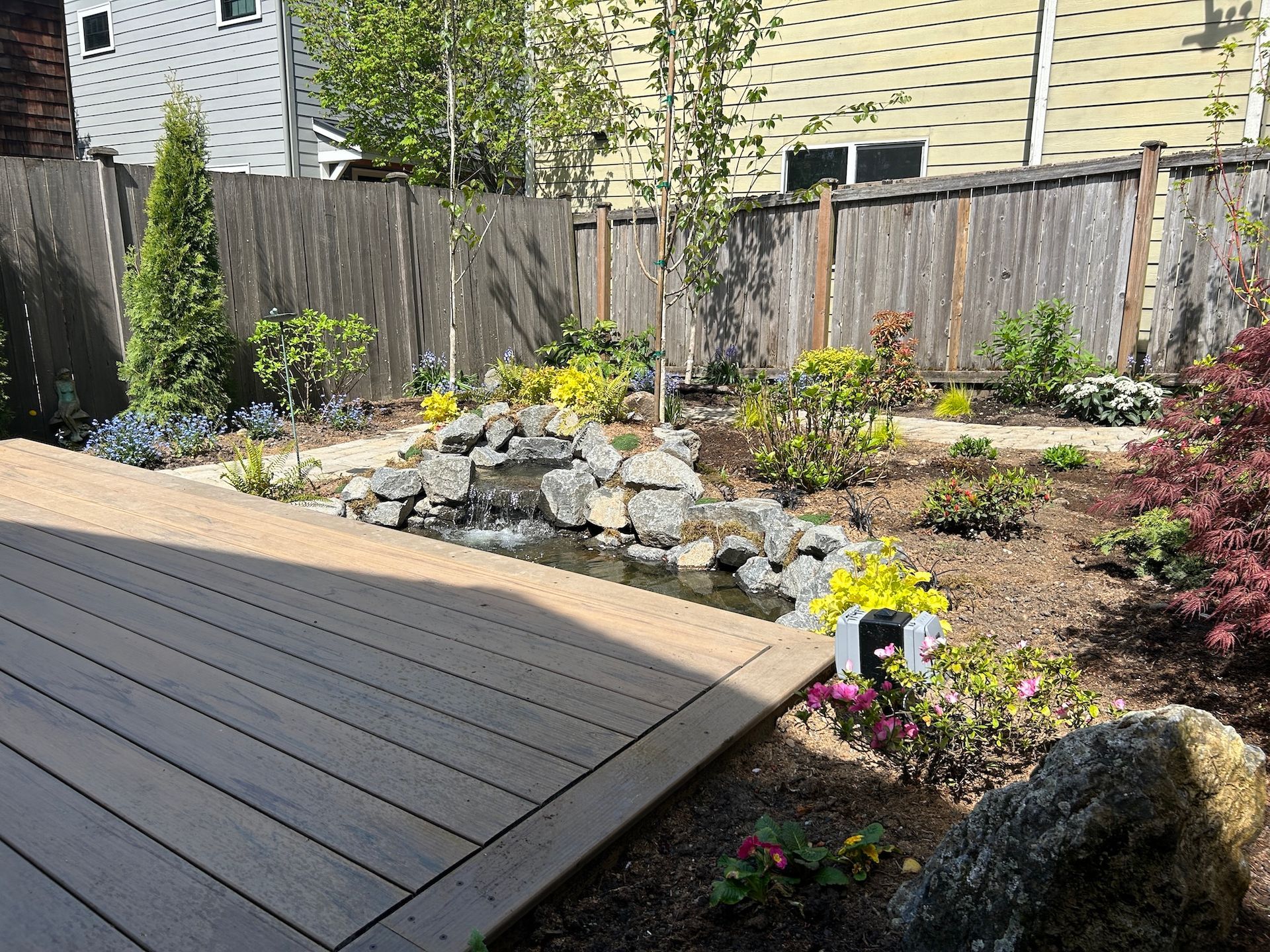 Backyard deck beside a rock garden with flowers and shrubs, bordered by a wooden fence.