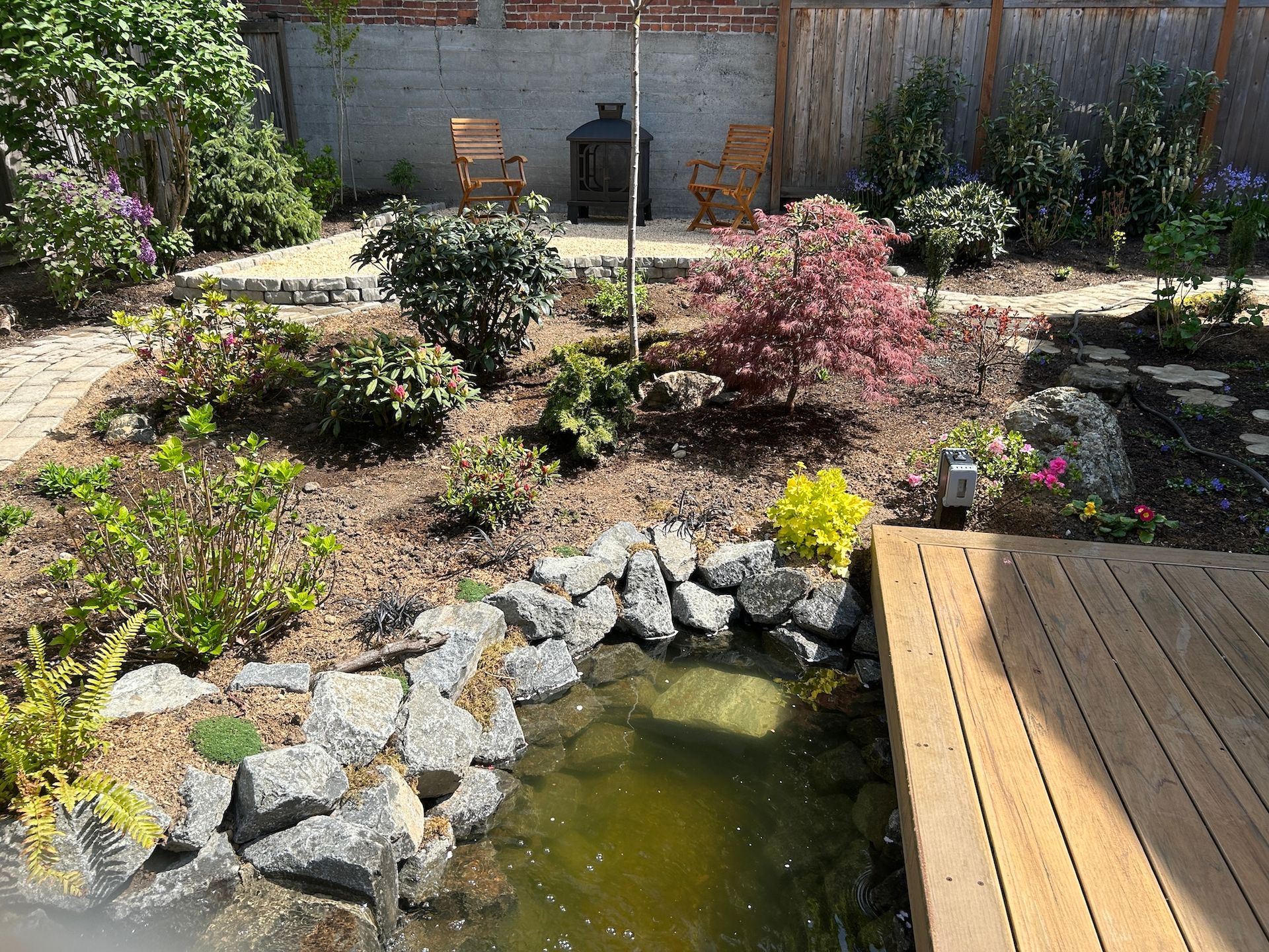 Garden pond beside a wooden deck, with rocks, shrubs, and a brick wall in the background.