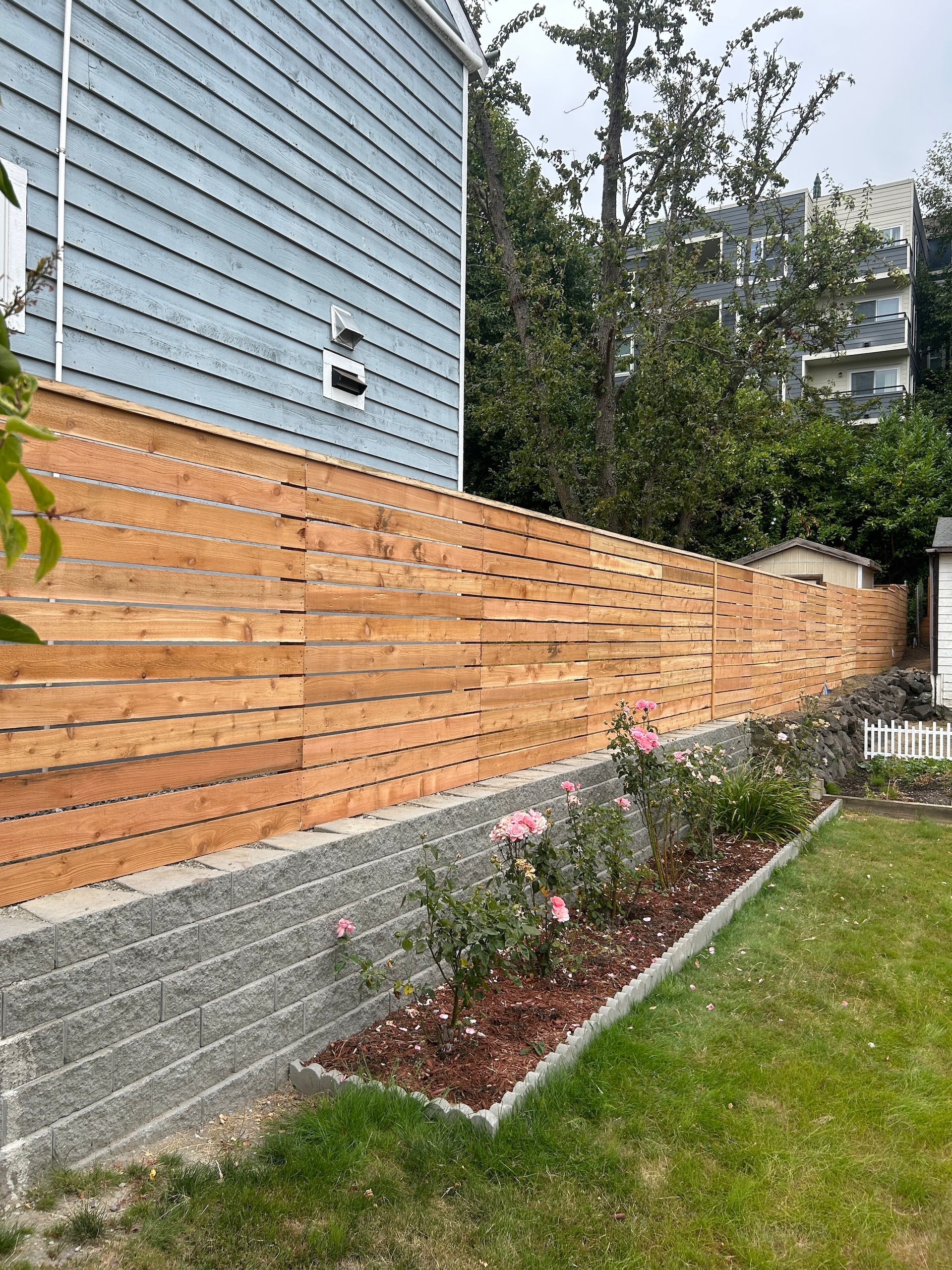 Backyard garden bed with pink flowers beside a wooden fence and blue-sided house