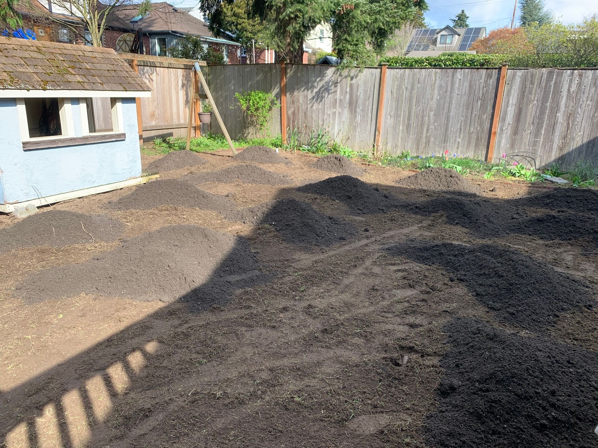 Backyard with large piles of dark soil beside a fence and a small shed in sunlight