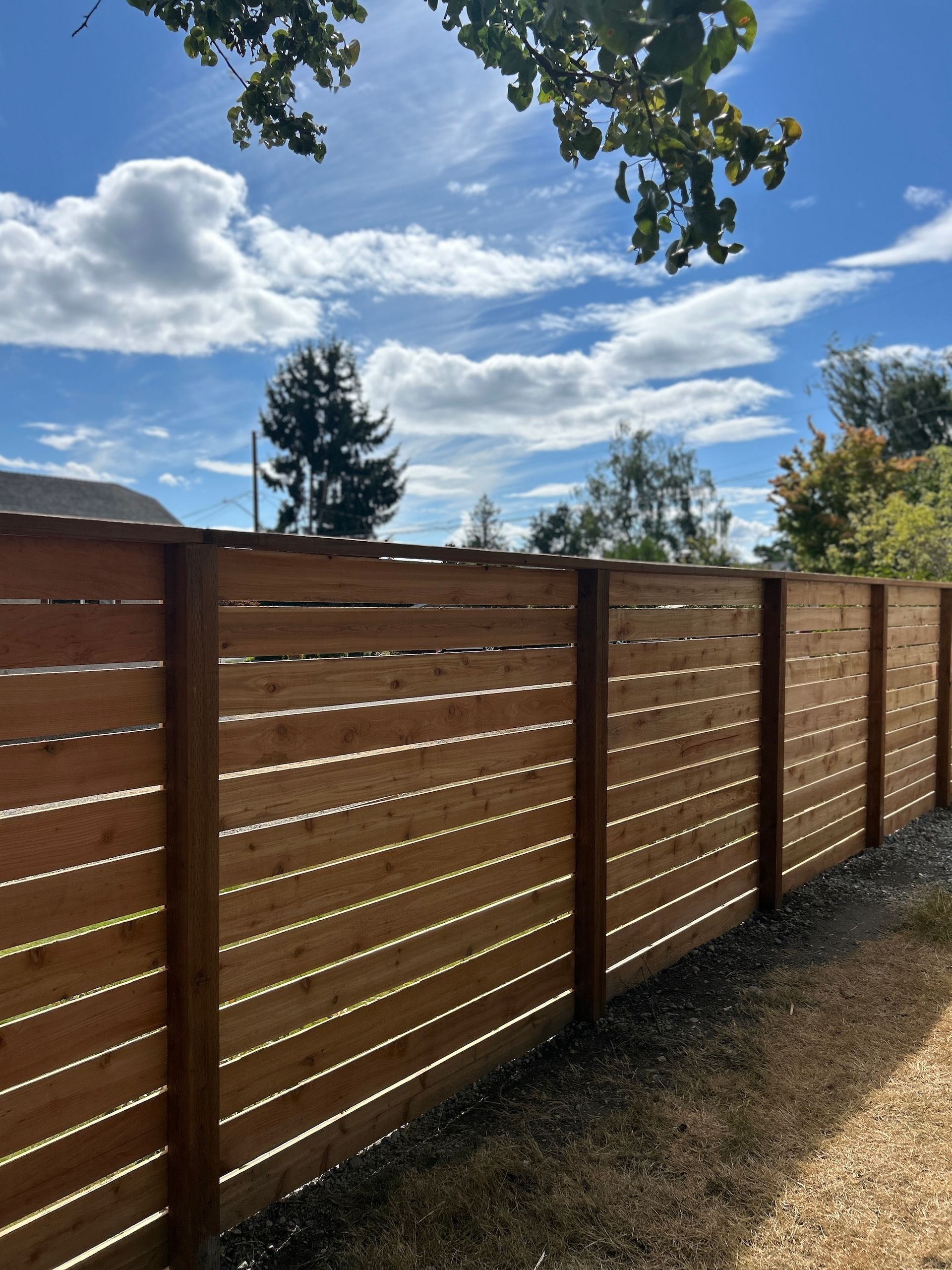 Wooden slat fence along a gravel yard under a bright blue sky with clouds.