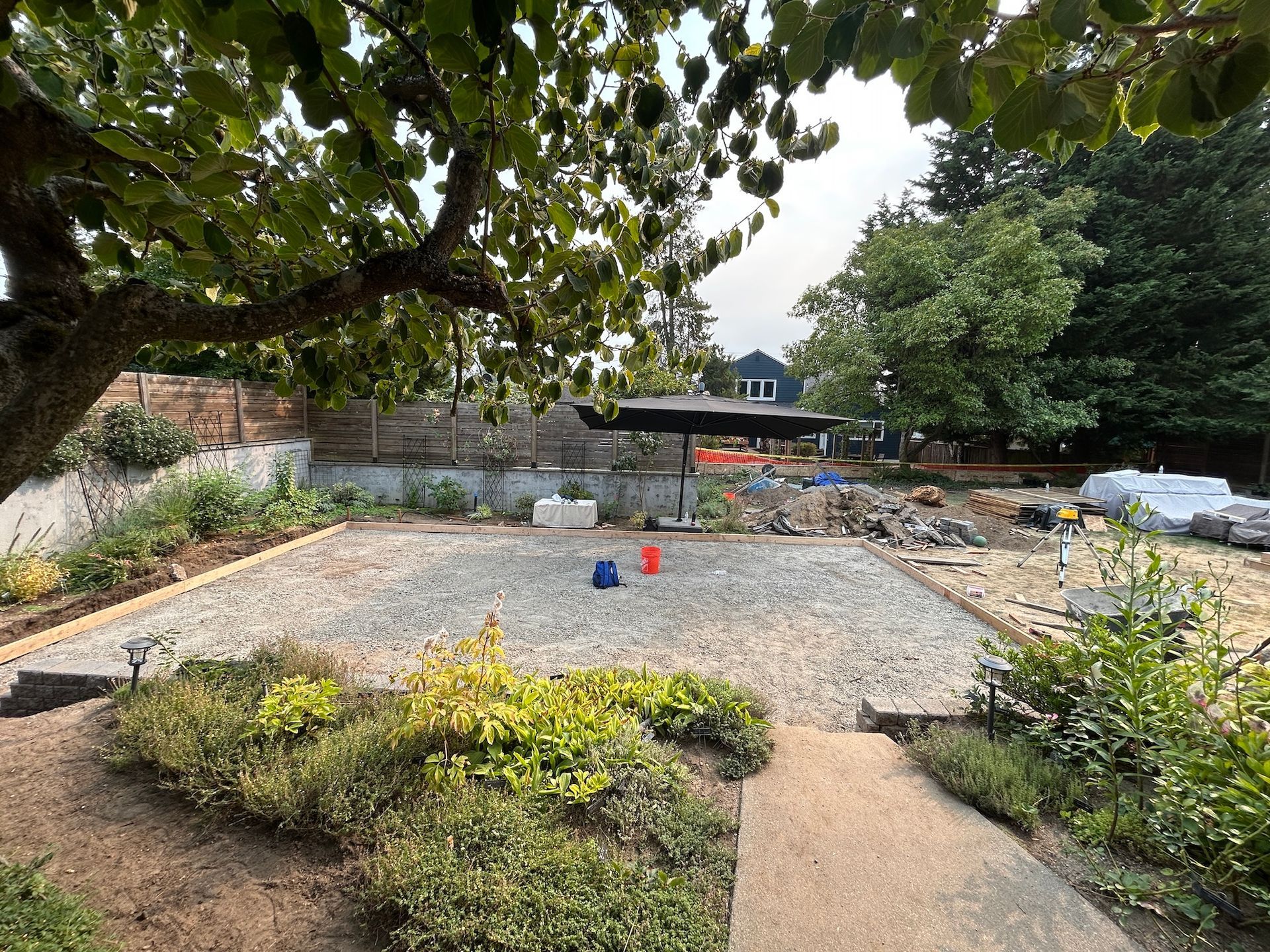 Gravel garden bed with small red and blue buckets beside a path, under a leafy tree.