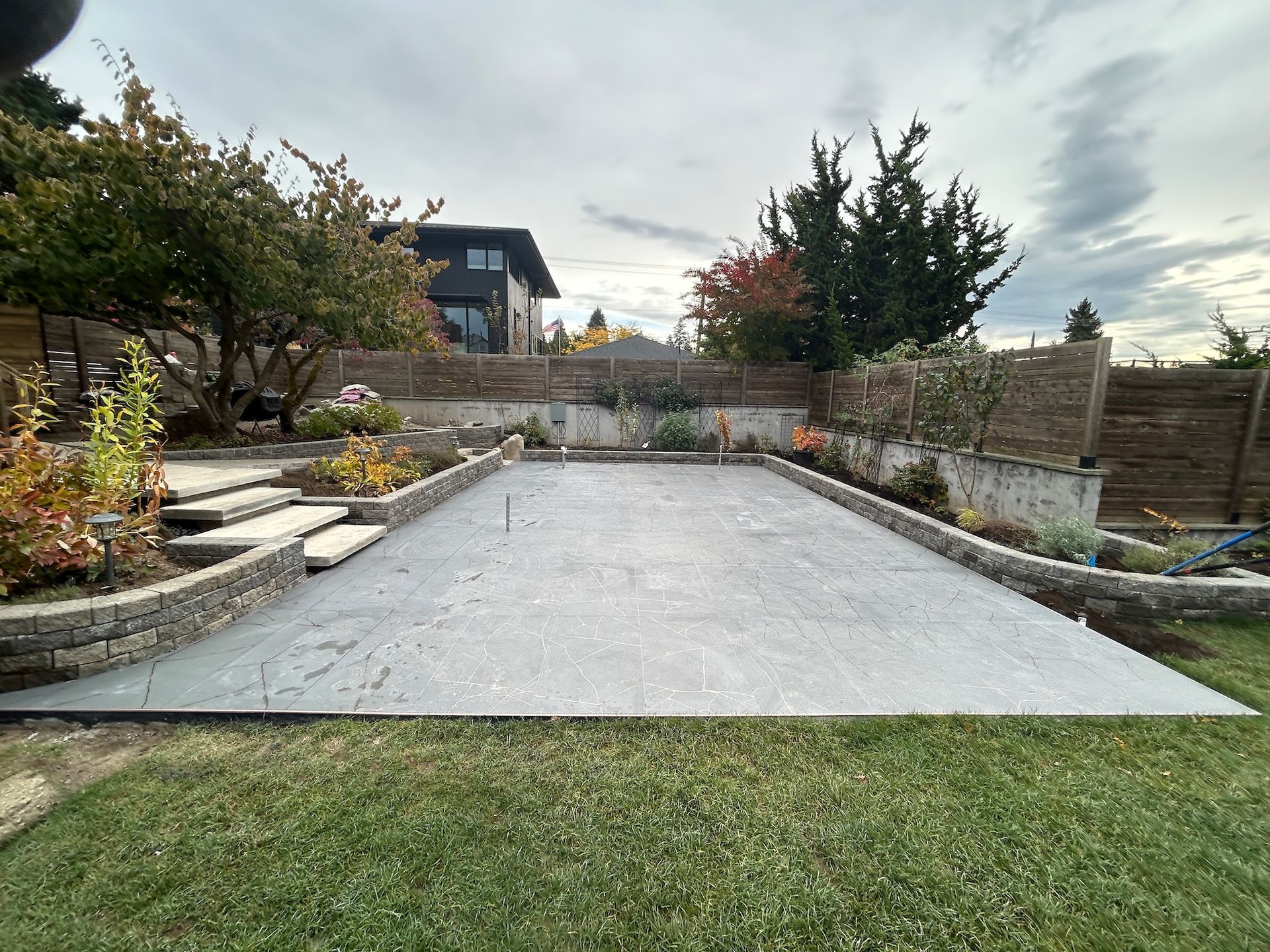 Backyard with a large concrete patio, lawn, stone steps, and raised garden beds under a cloudy sky