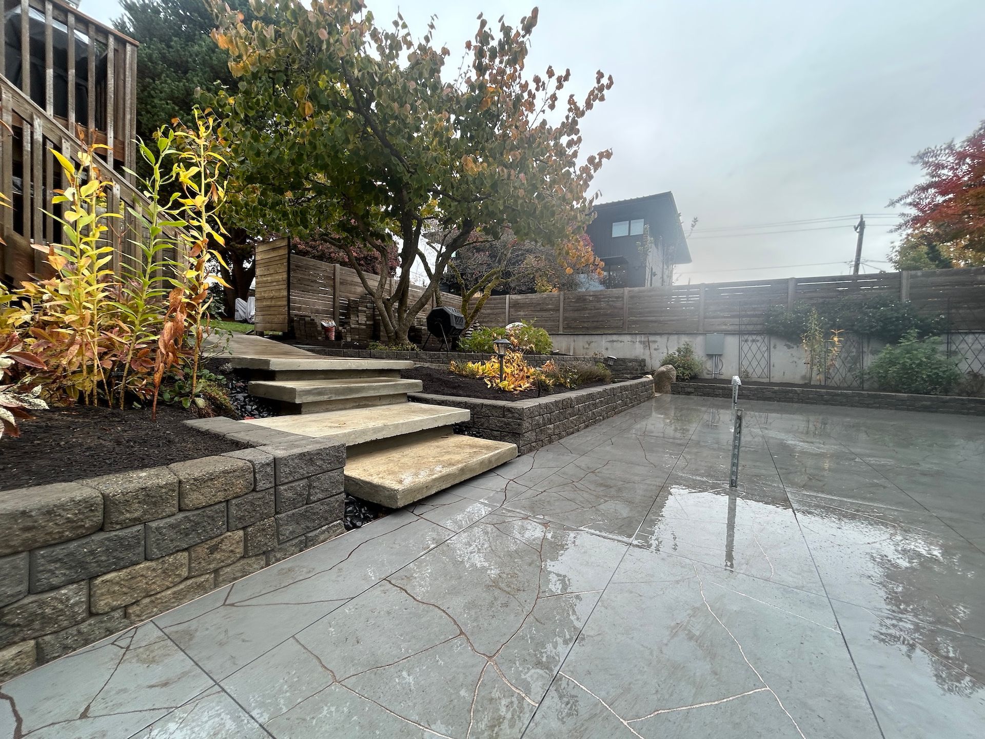 Stone patio with steps, planters, and a wet reflective surface in a landscaped backyard