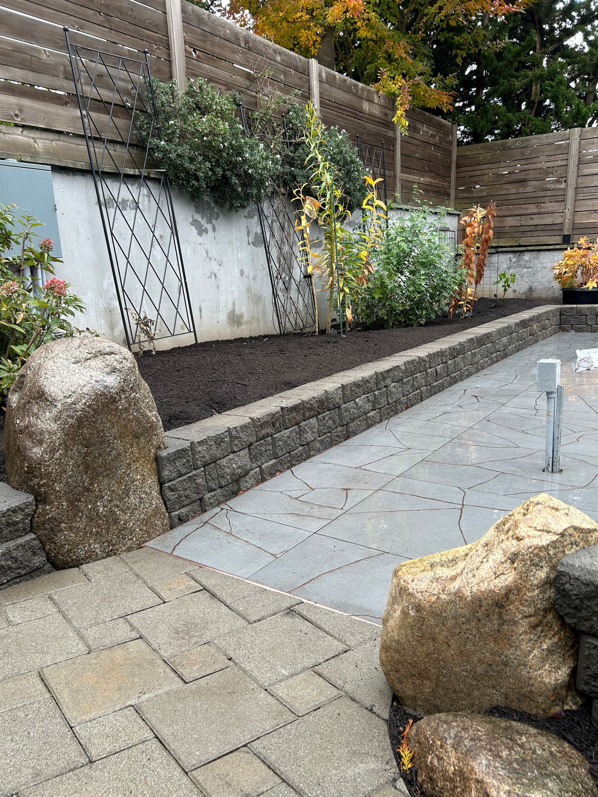 Outdoor stone patio with large boulders, raised planter, and wooden fence in a landscaped yard
