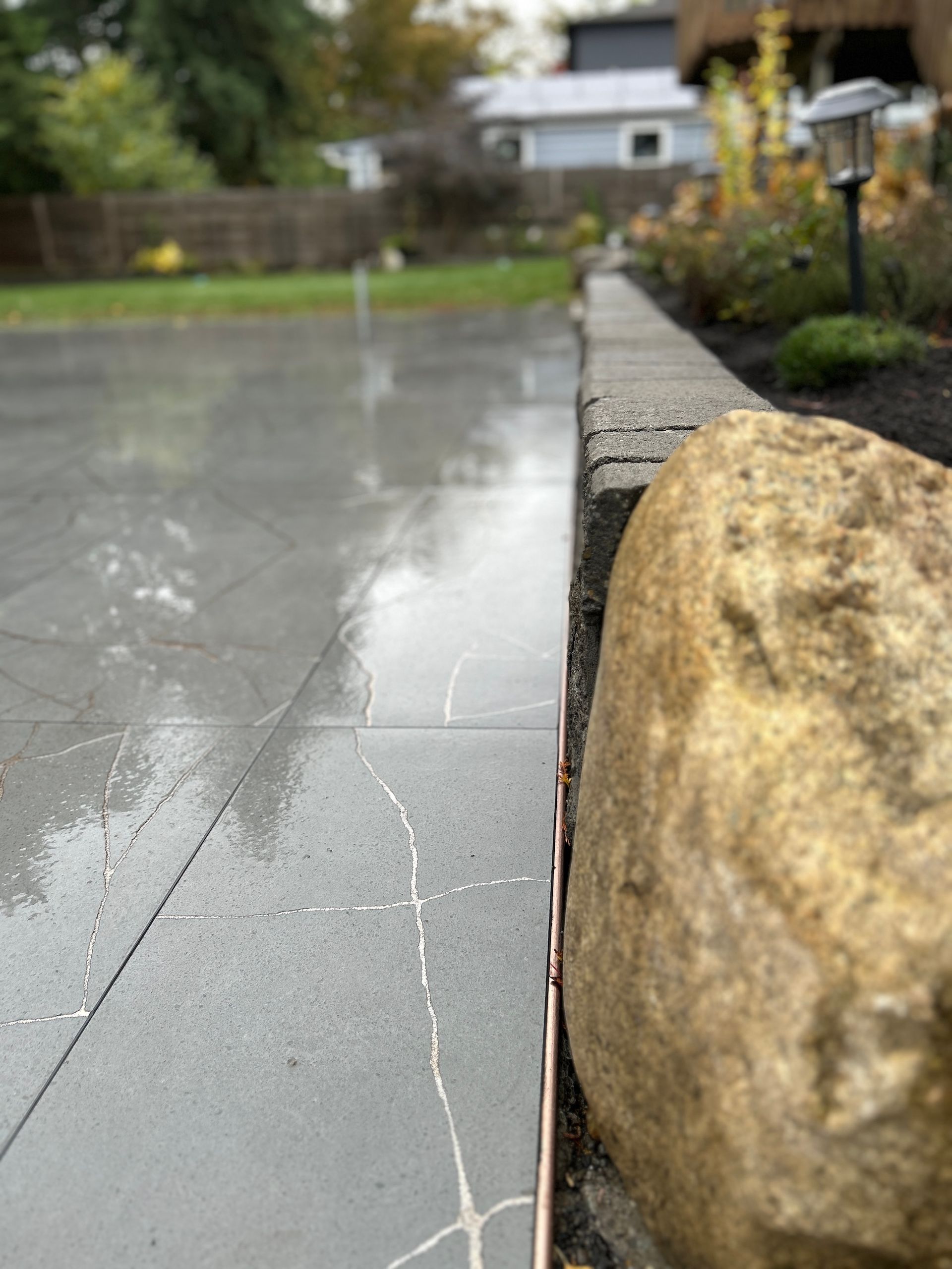 Rain-soaked stone patio beside a garden path with a large rock in the foreground