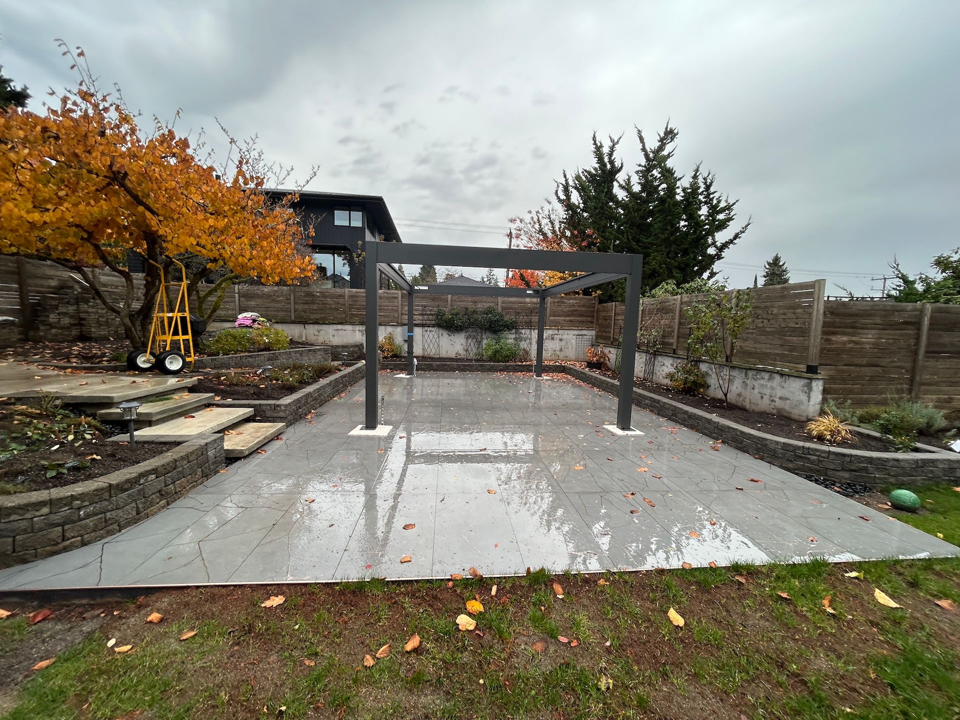 Backyard patio with a wet concrete slab, metal pergola frame, autumn tree, and excavator on the left