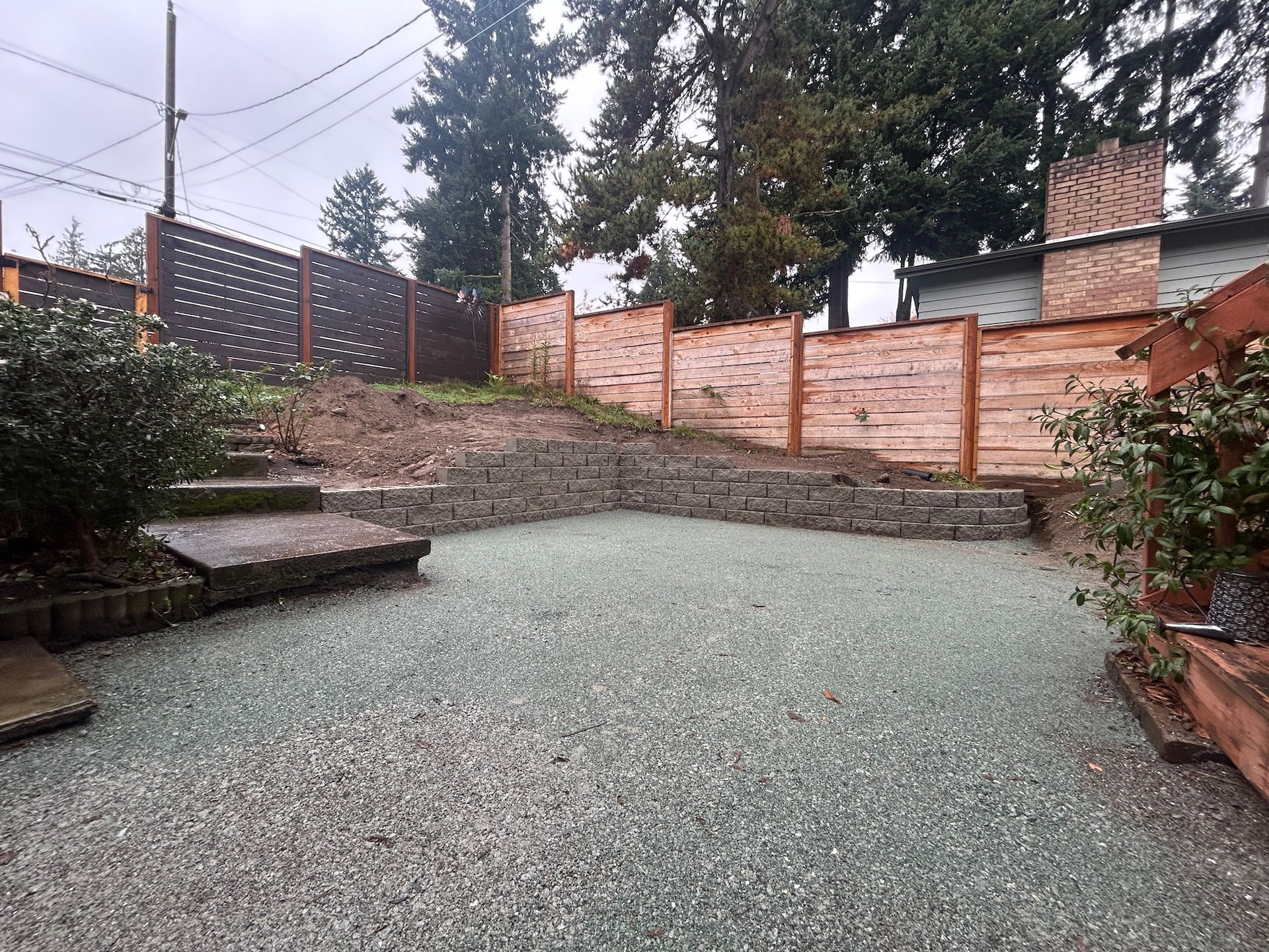 Gravel backyard patio bordered by wood and brick fencing, with steps and shrubs on the sides.