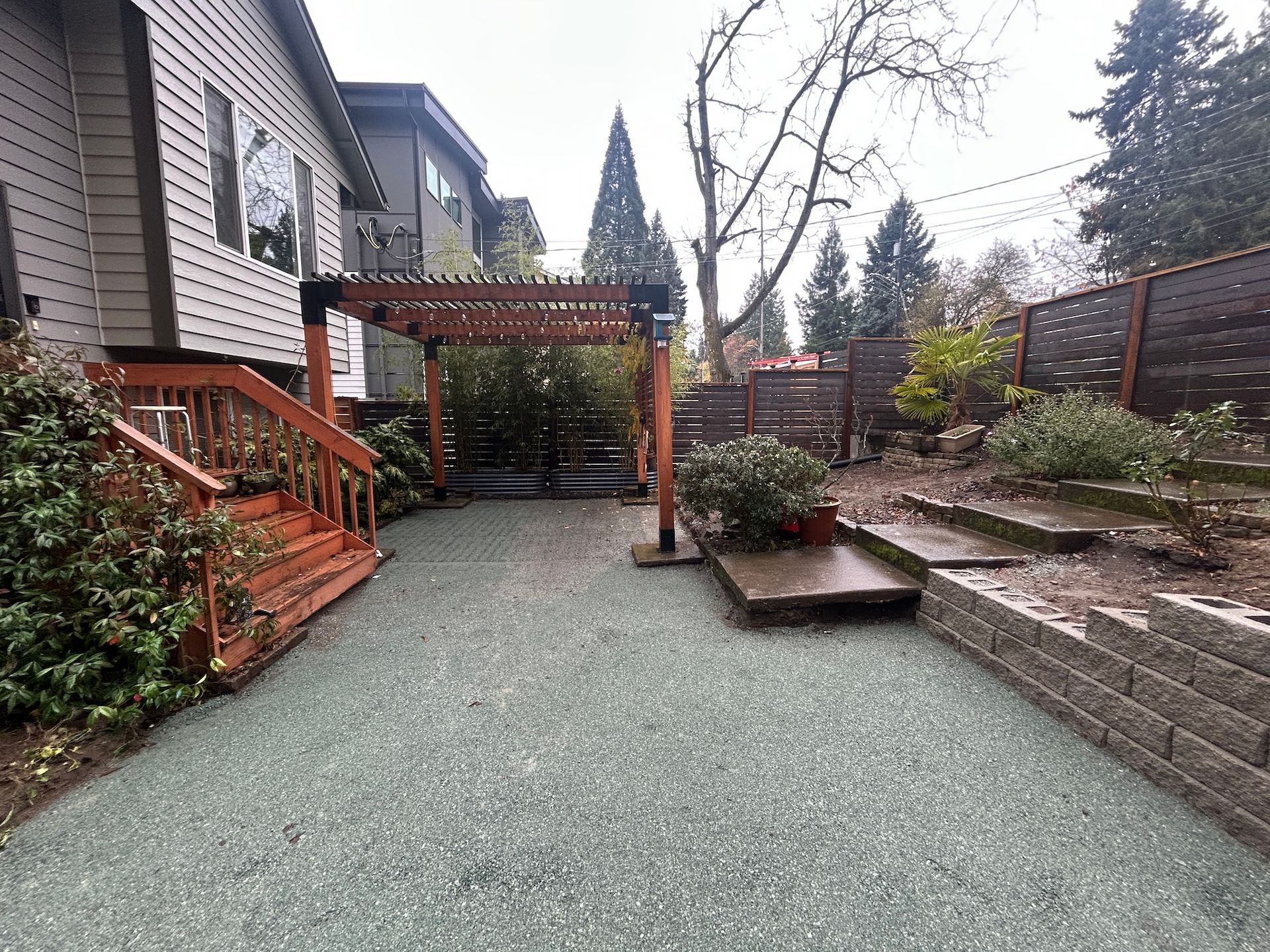 Gravel backyard patio with wooden deck stairs, pergola, raised planters, and retaining wall under bare trees